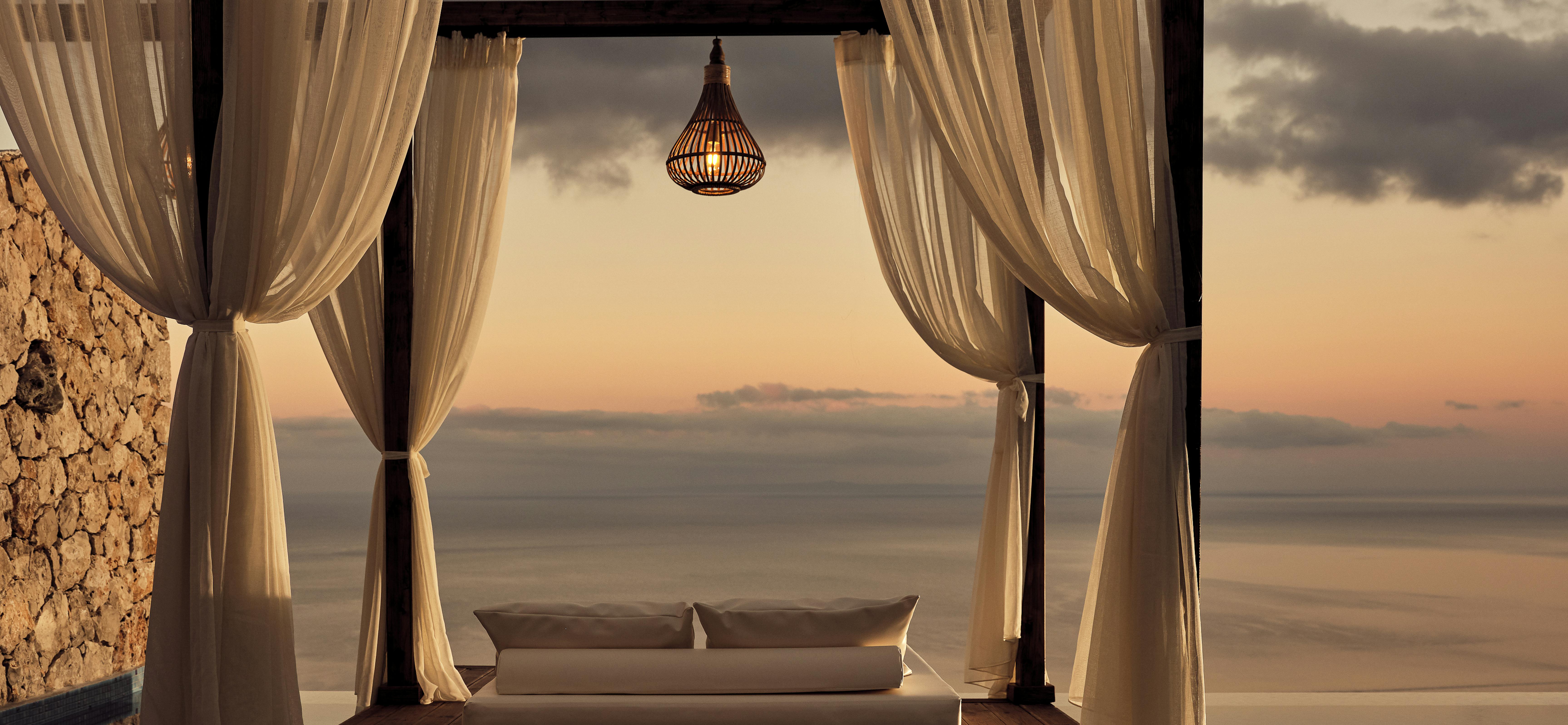 Poolside cabana with white cushioned daybed draped in sheer curtains, woven pendant light, and ocean sunset backdrop.