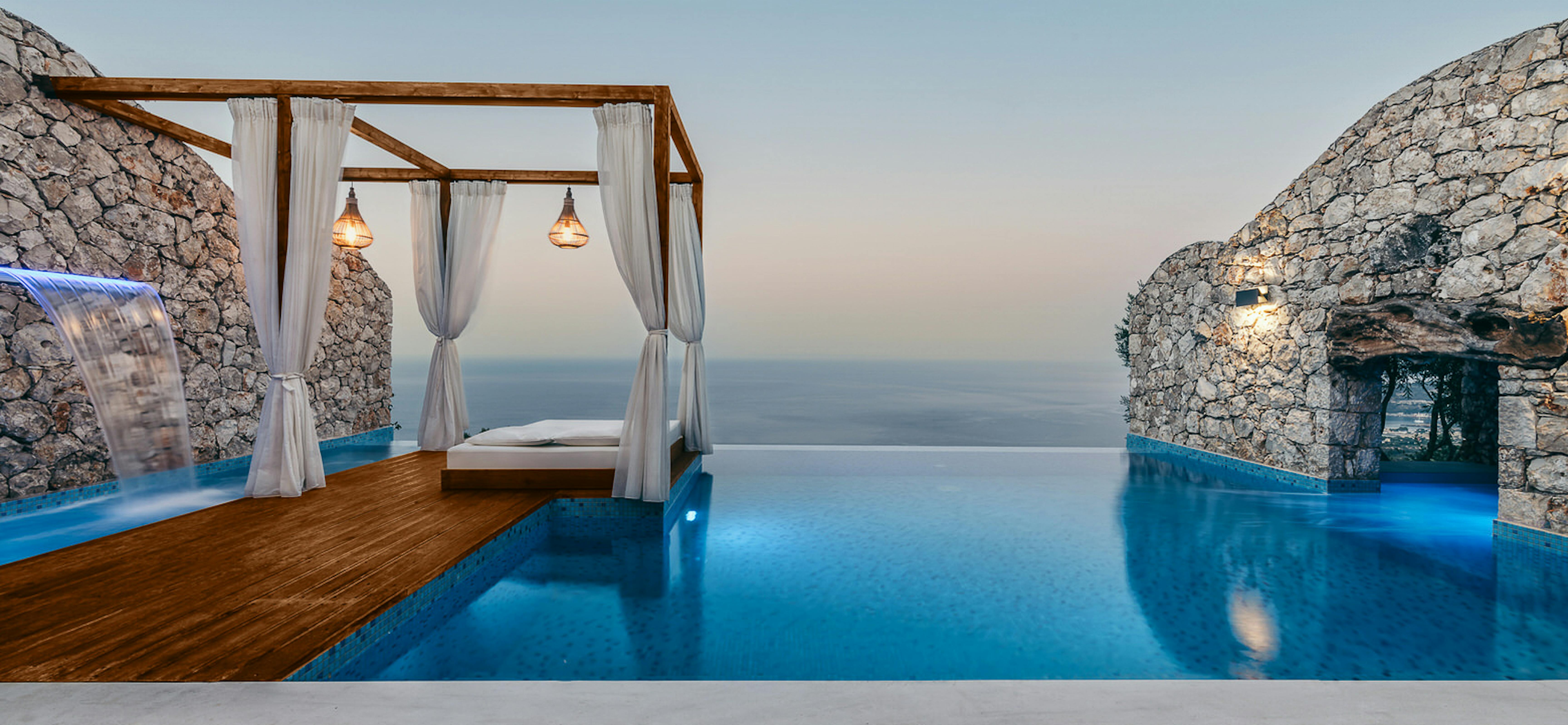 Twilight view of illuminated infinity pool with curtained cabana platform set between dramatic stone walls and ocean backdrop.