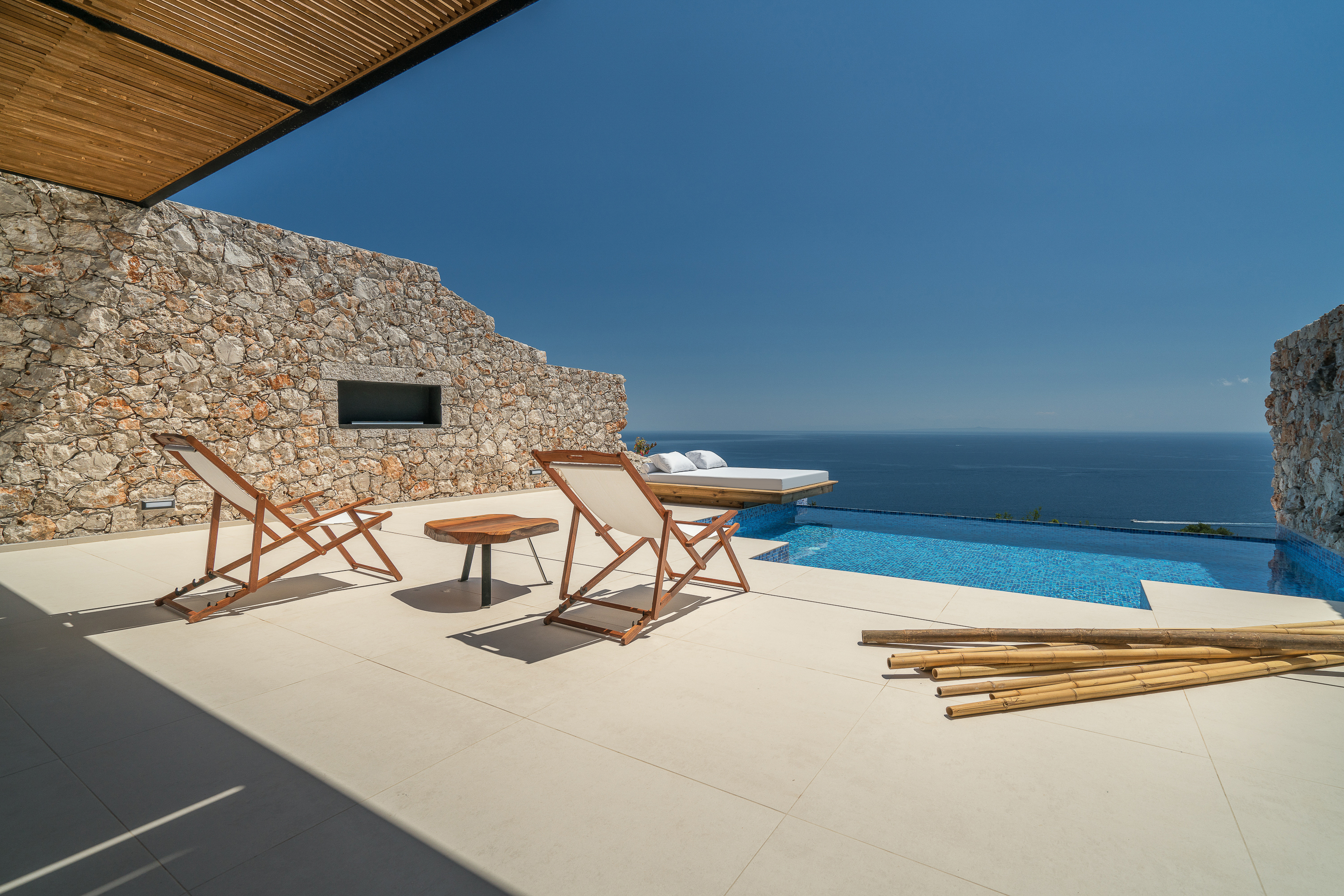 Covered terrace with wooden folding chairs and side table overlooking infinity pool, white daybed platform, and endless ocean.