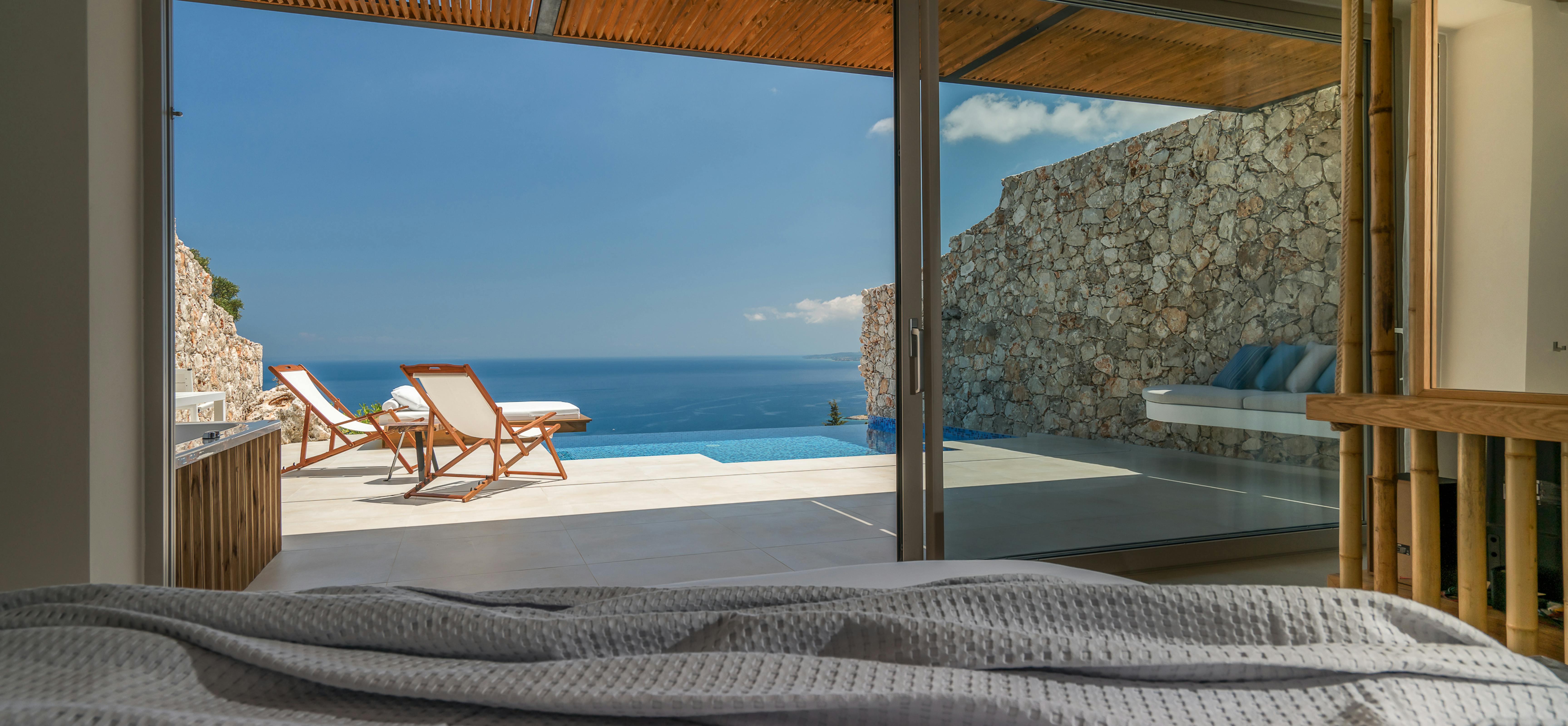 Bedroom view through sliding glass doors to infinity pool terrace with wooden deck chairs and endless ocean horizon beyond.