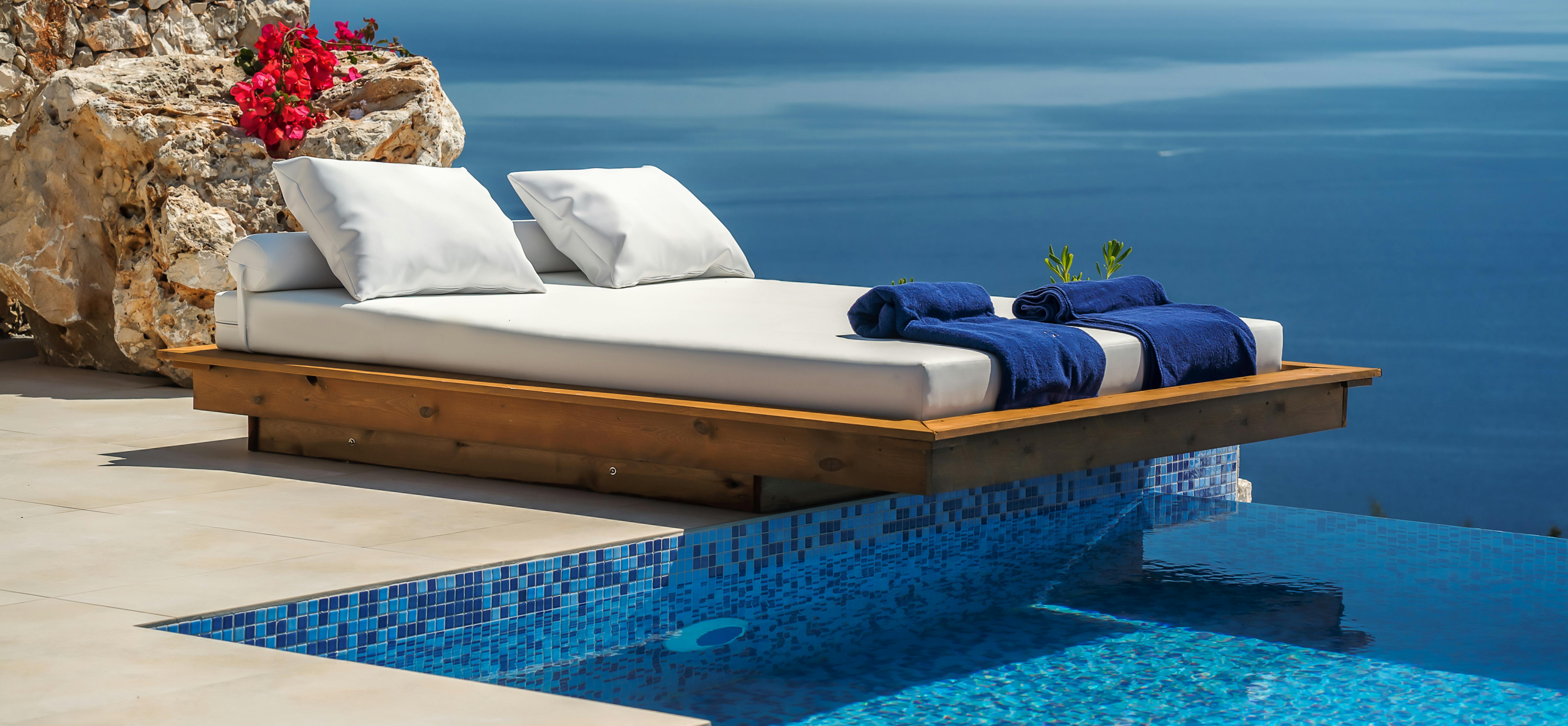 Infinity pool platform with white cushioned daybed, navy towels, and red bougainvillea against weathered stone wall overlooking sea.