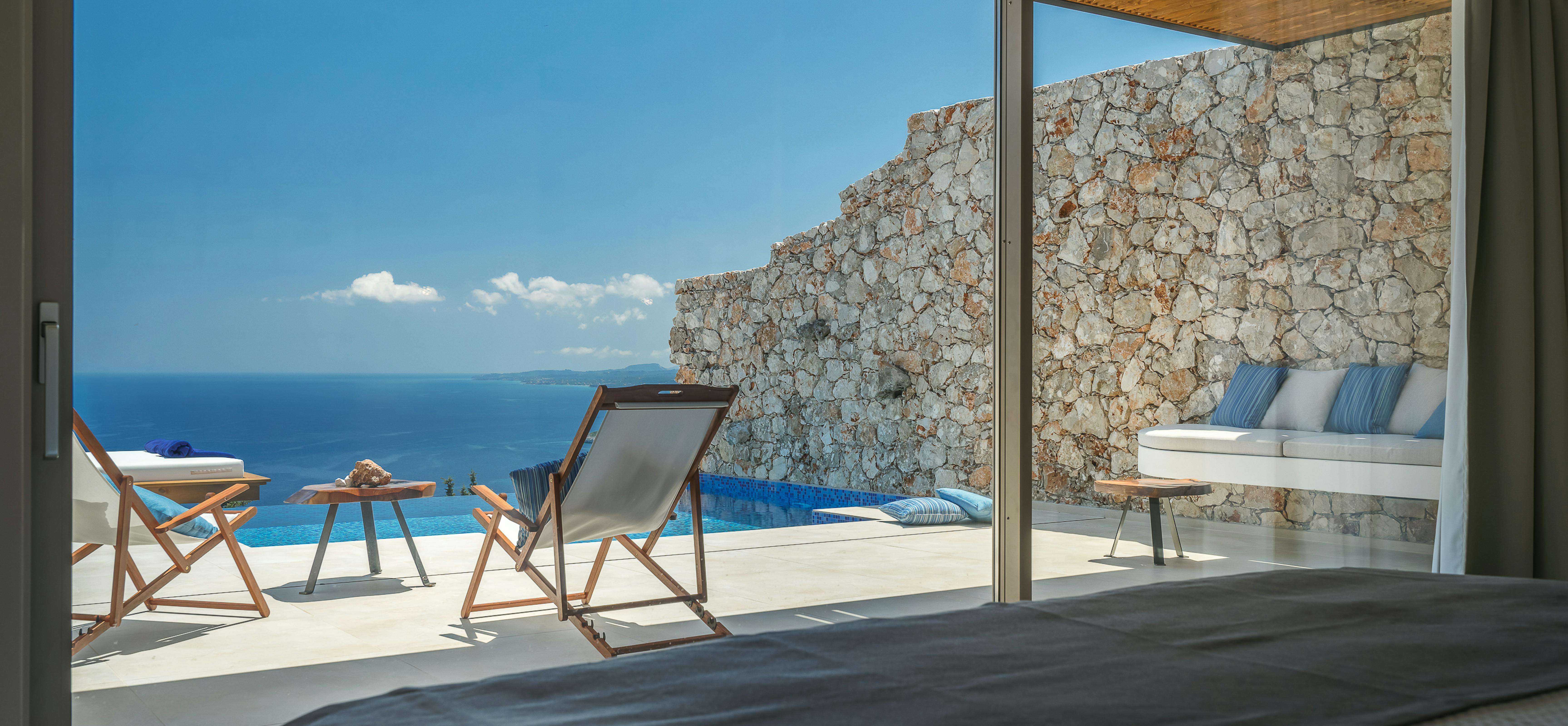 View from bedroom through open sliding doors to pool terrace with wooden loungers, stone walls, and panoramic ocean views.