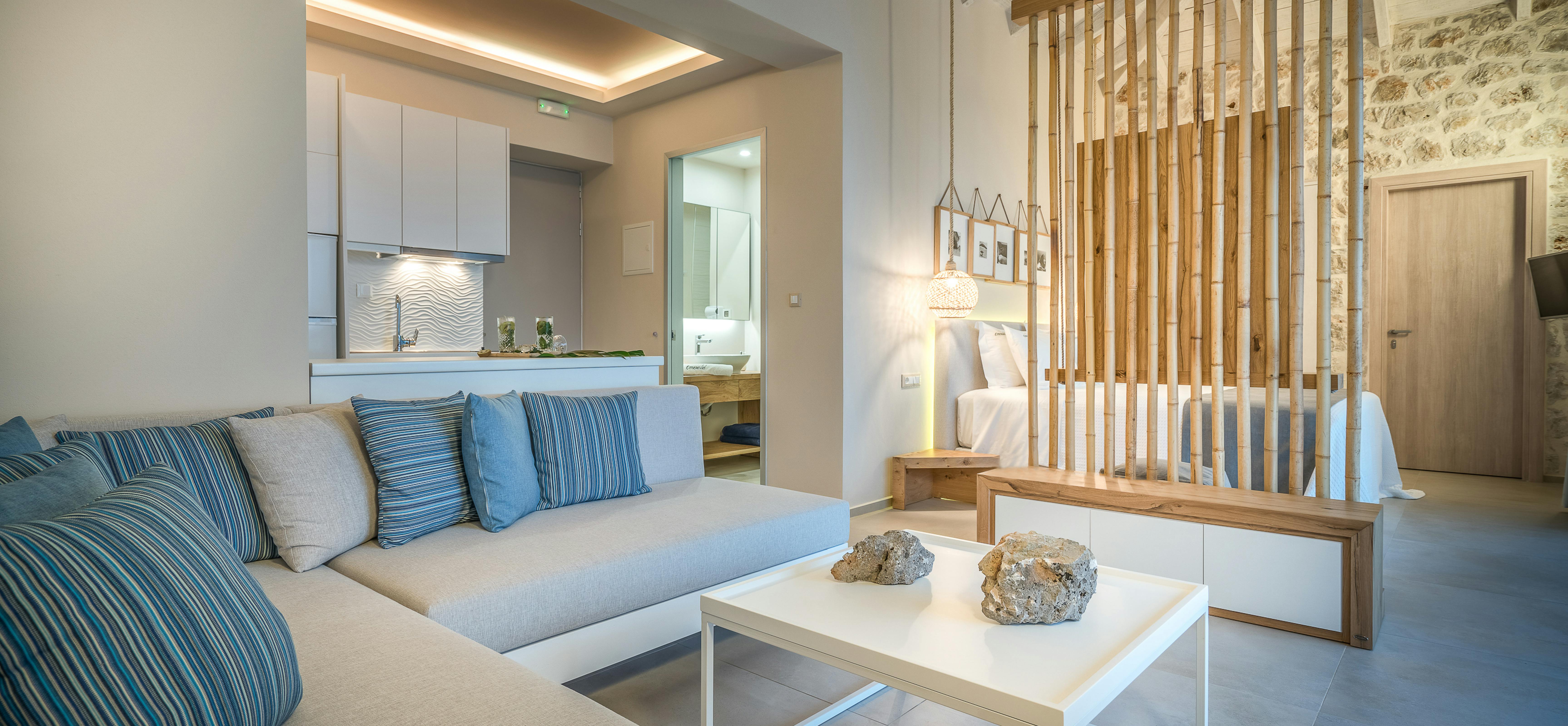 Open-plan living space with blue striped sectional sofa, vertical wood slat divider, exposed beam ceiling, and kitchen with ocean view.