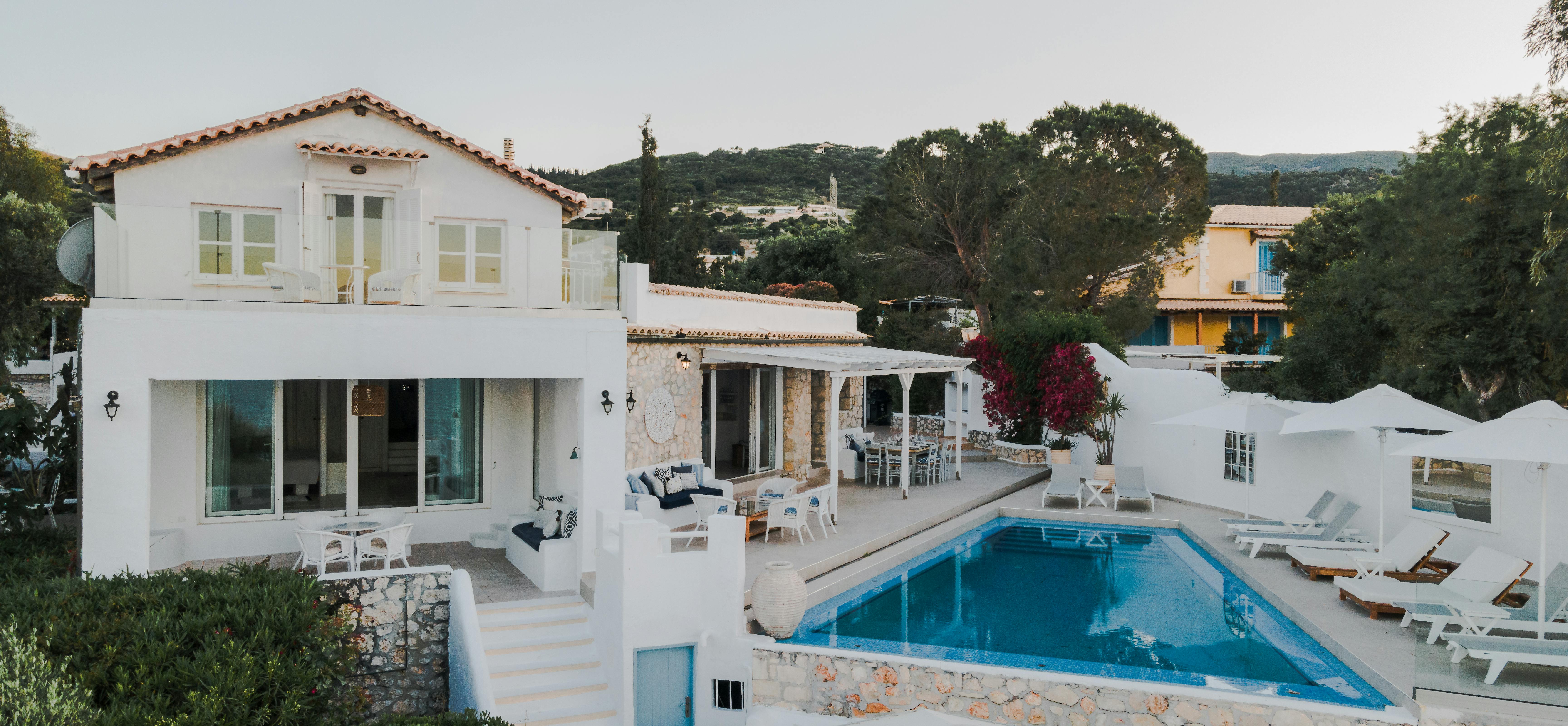 White Mediterranean villa at dusk with multi-level terraces, blue-tiled swimming pool, white sun loungers, and lush hillside setting.