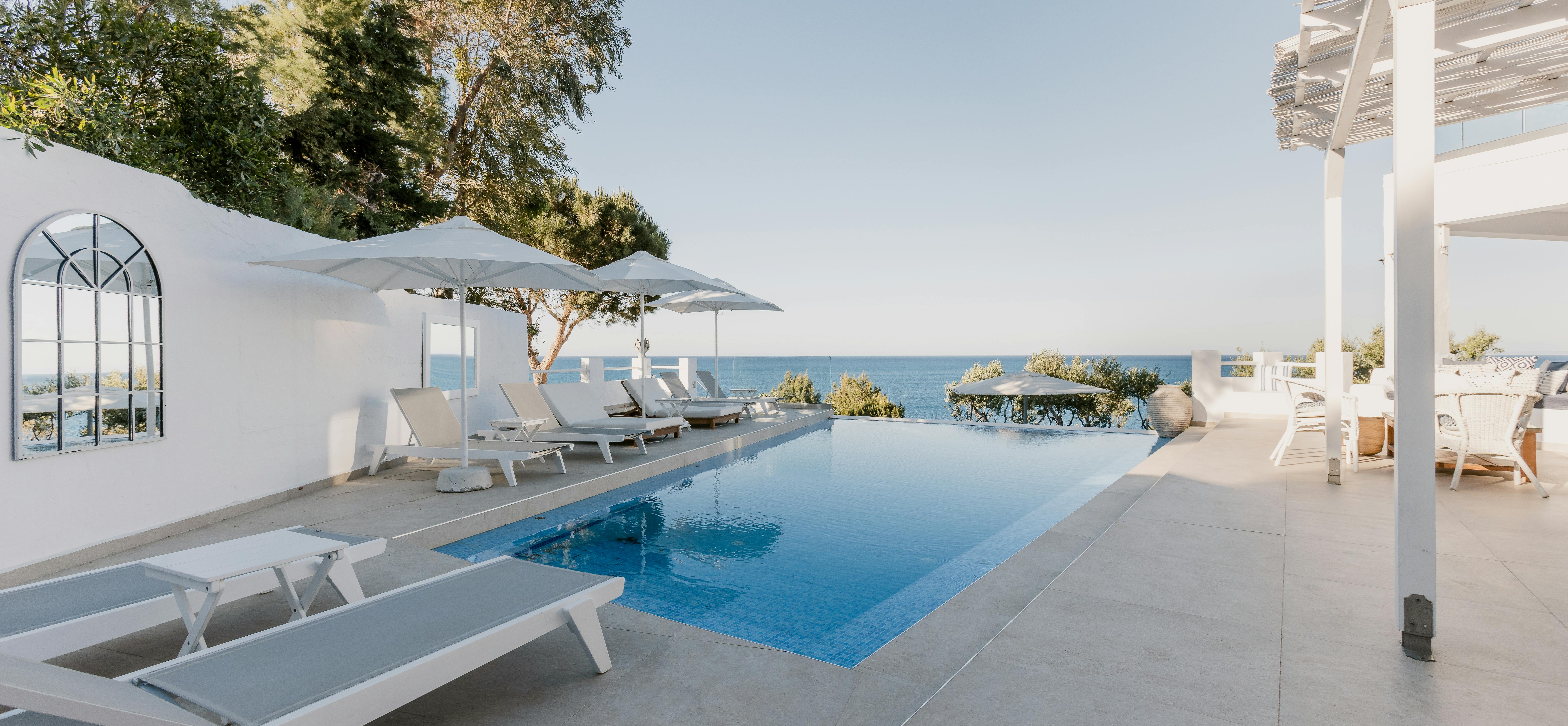 Minimalist pool terrace with white loungers, arched window detail in wall, and serene ocean views framed by Mediterranean greenery.
