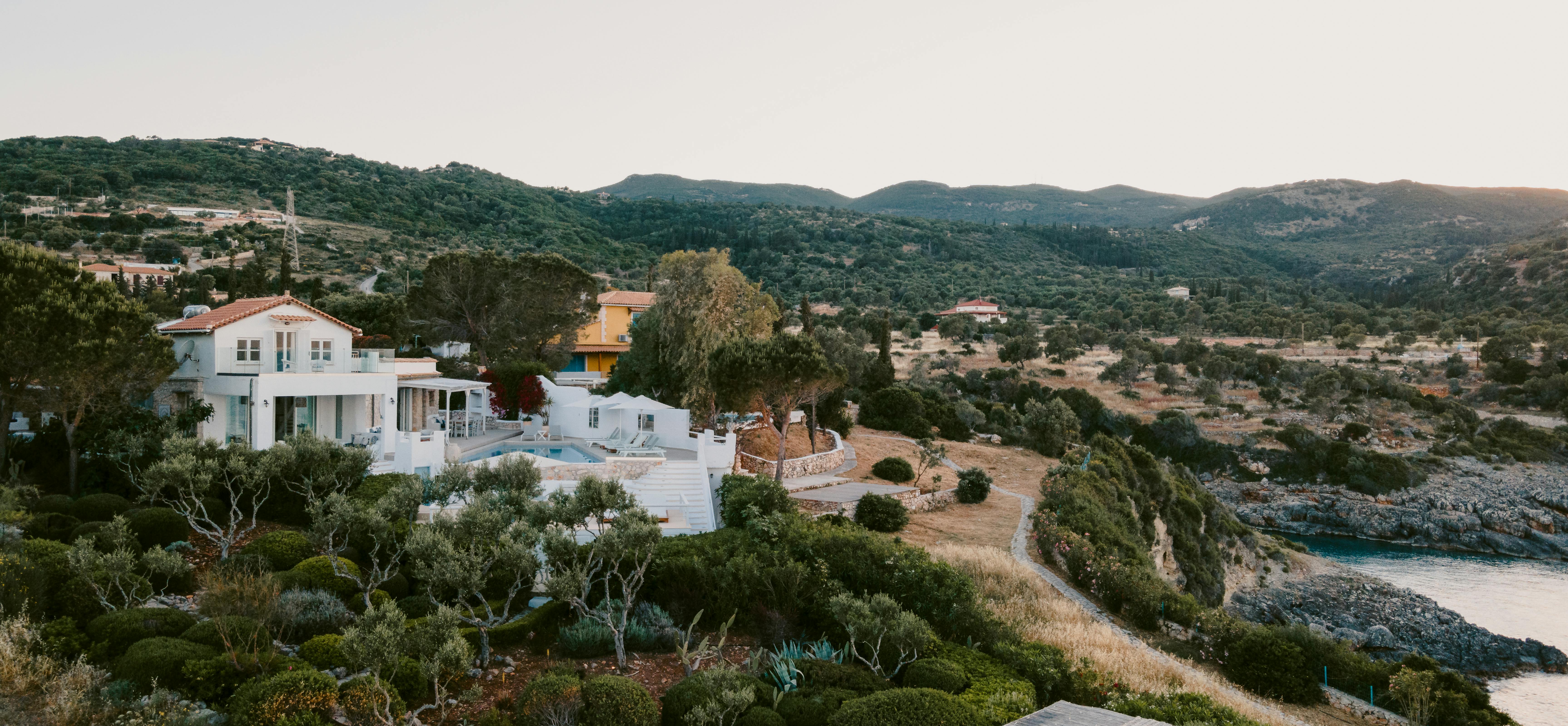 Coastal hillside villa overlooking rocky cove at sunset, surrounded by Mediterranean gardens, olive trees, and stone terracing.