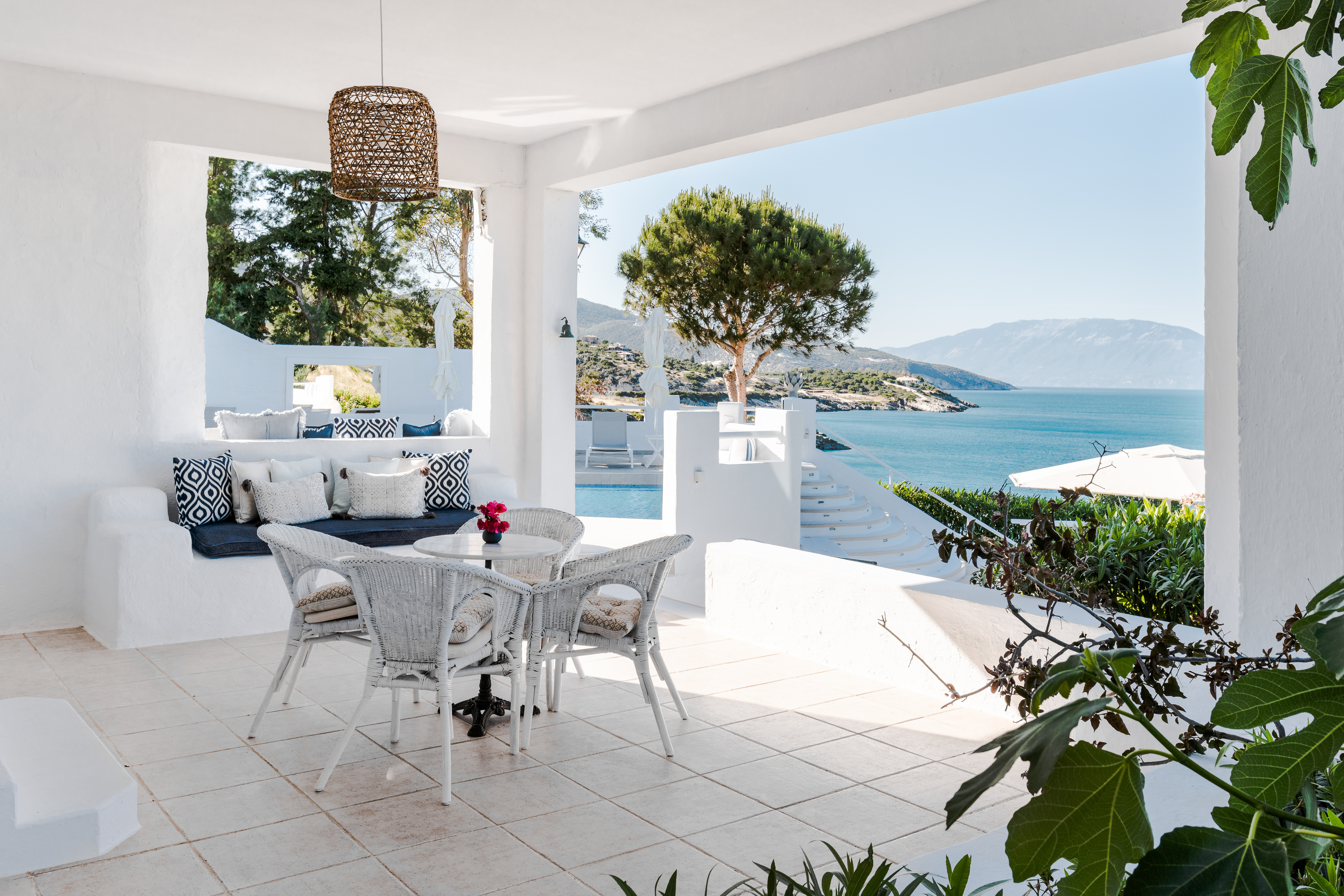 Covered white terrace with wicker dining set, built-in banquette with geometric pillows, and panoramic coastal views through arched openings.