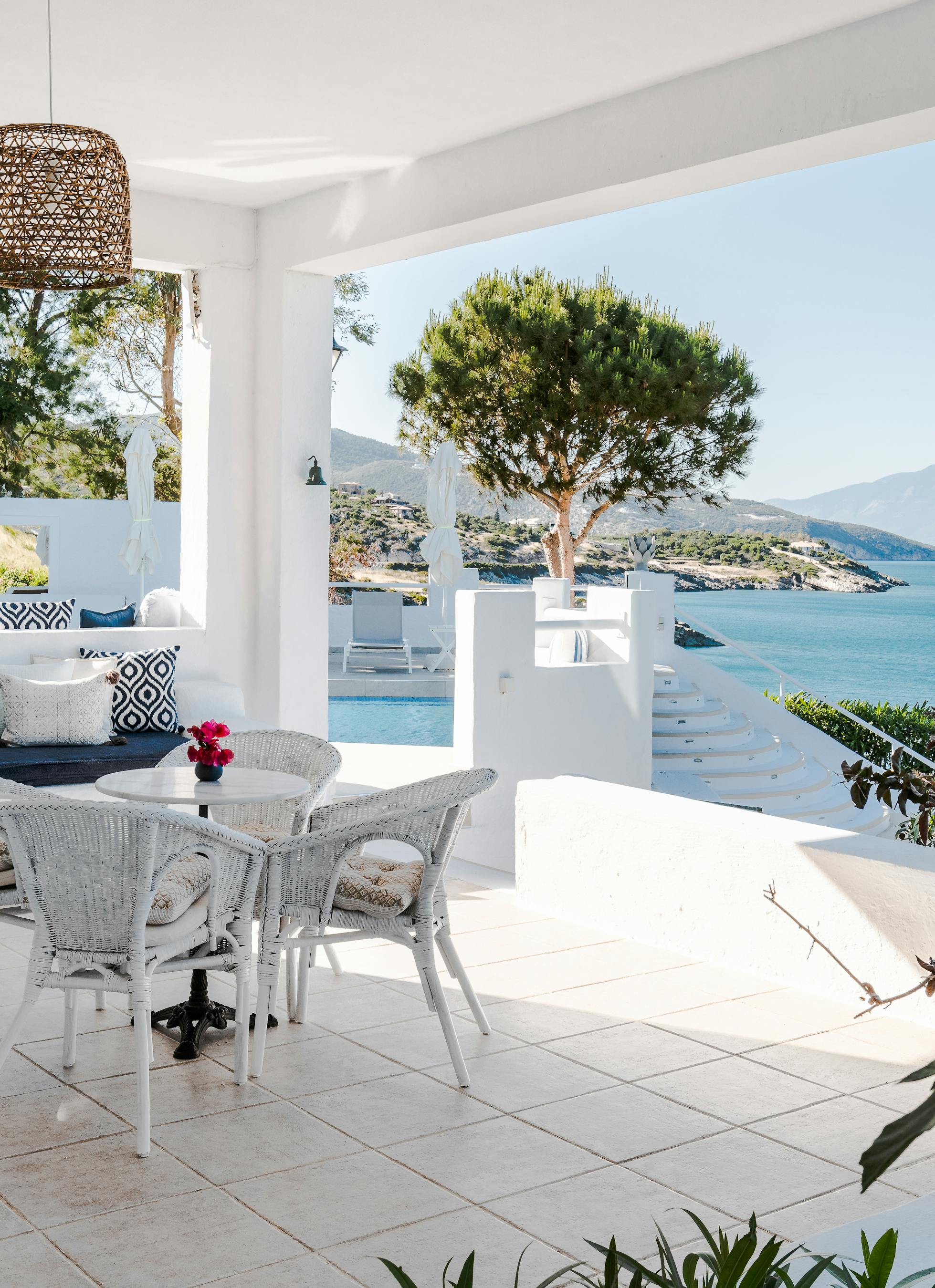 Covered white terrace with wicker dining set, built-in banquette with geometric pillows, and panoramic coastal views through arched openings.