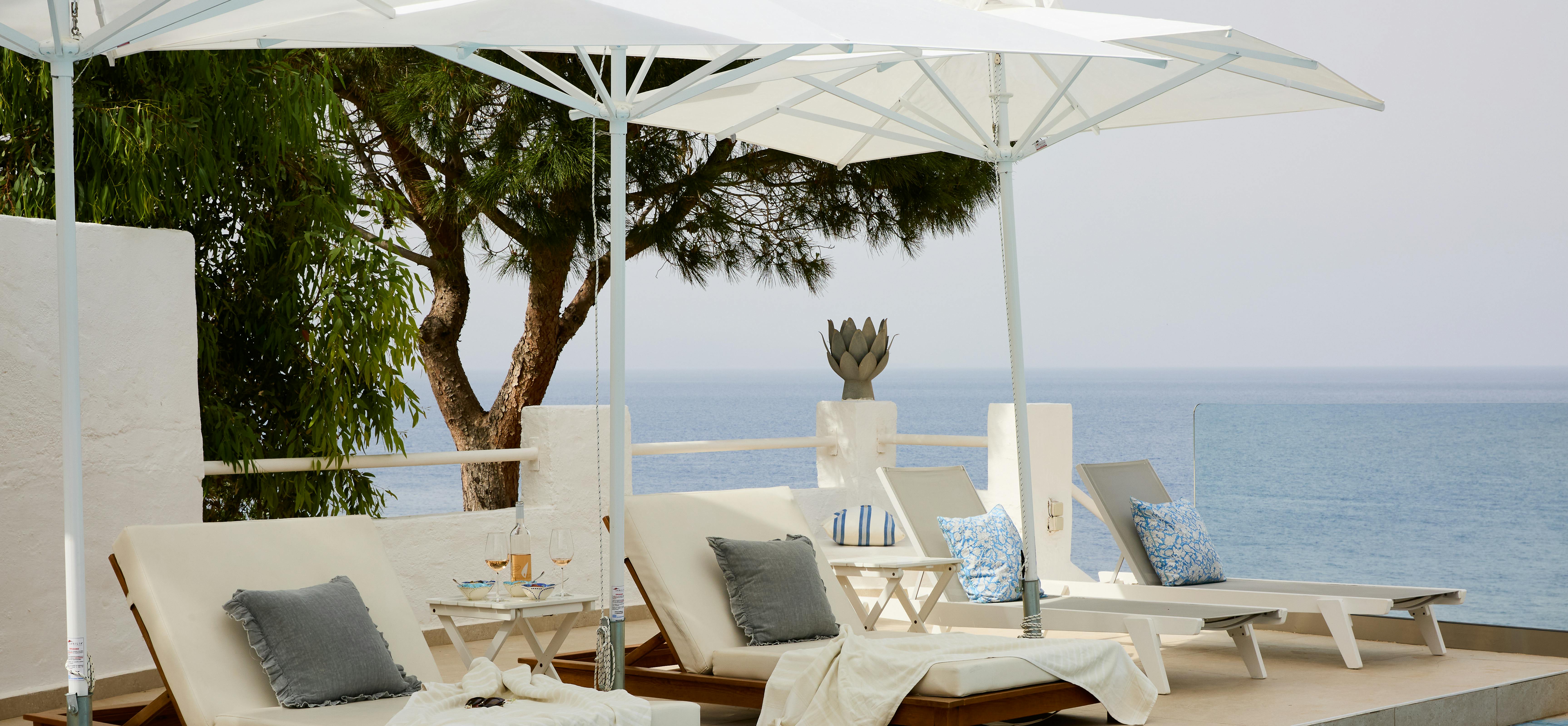 Poolside terrace with white umbrella shading wooden sun loungers with grey cushions, white walls, pine tree, and endless ocean horizon beyond.