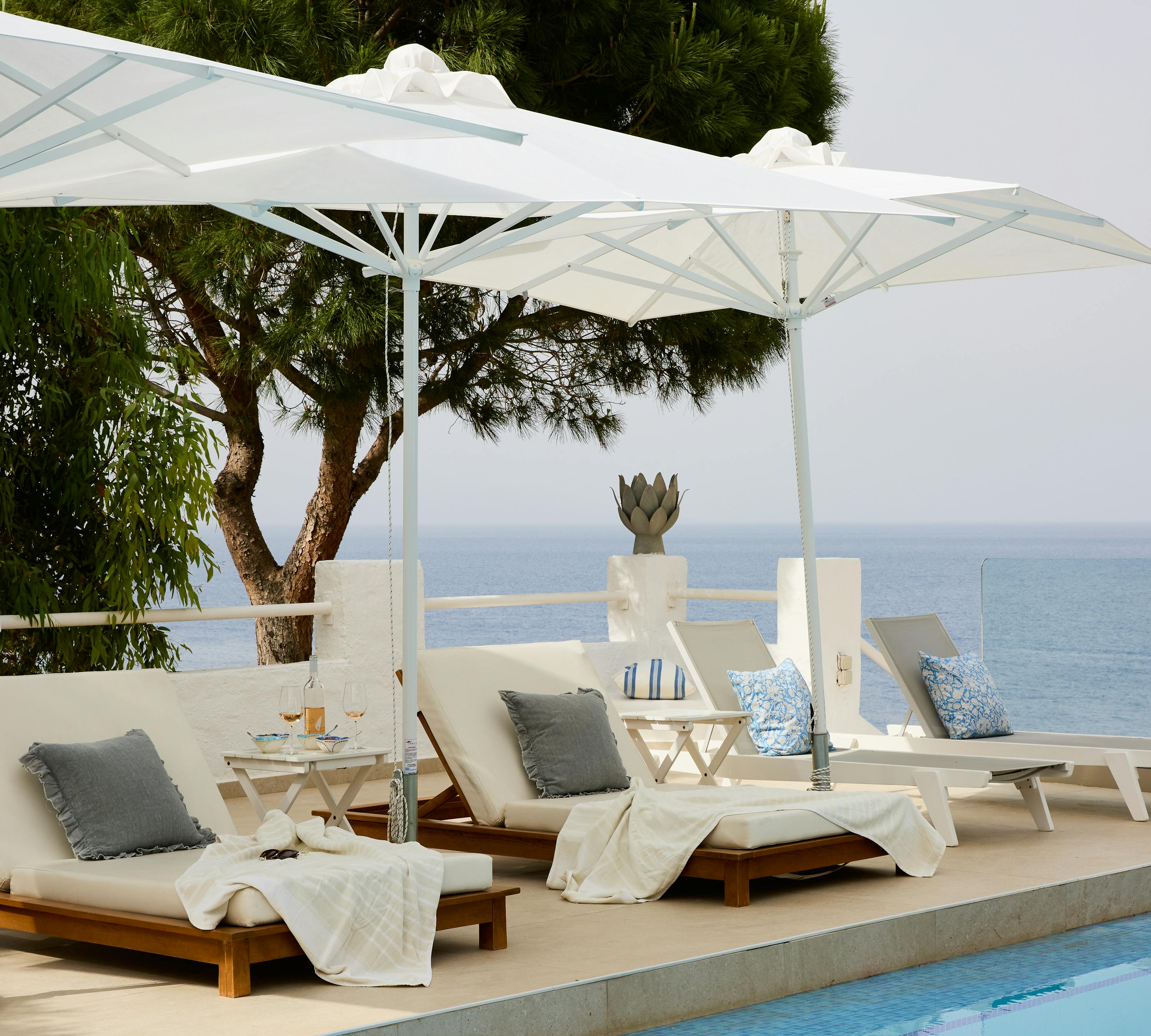 Poolside terrace with white umbrella shading wooden sun loungers with grey cushions, white walls, pine tree, and endless ocean horizon beyond.