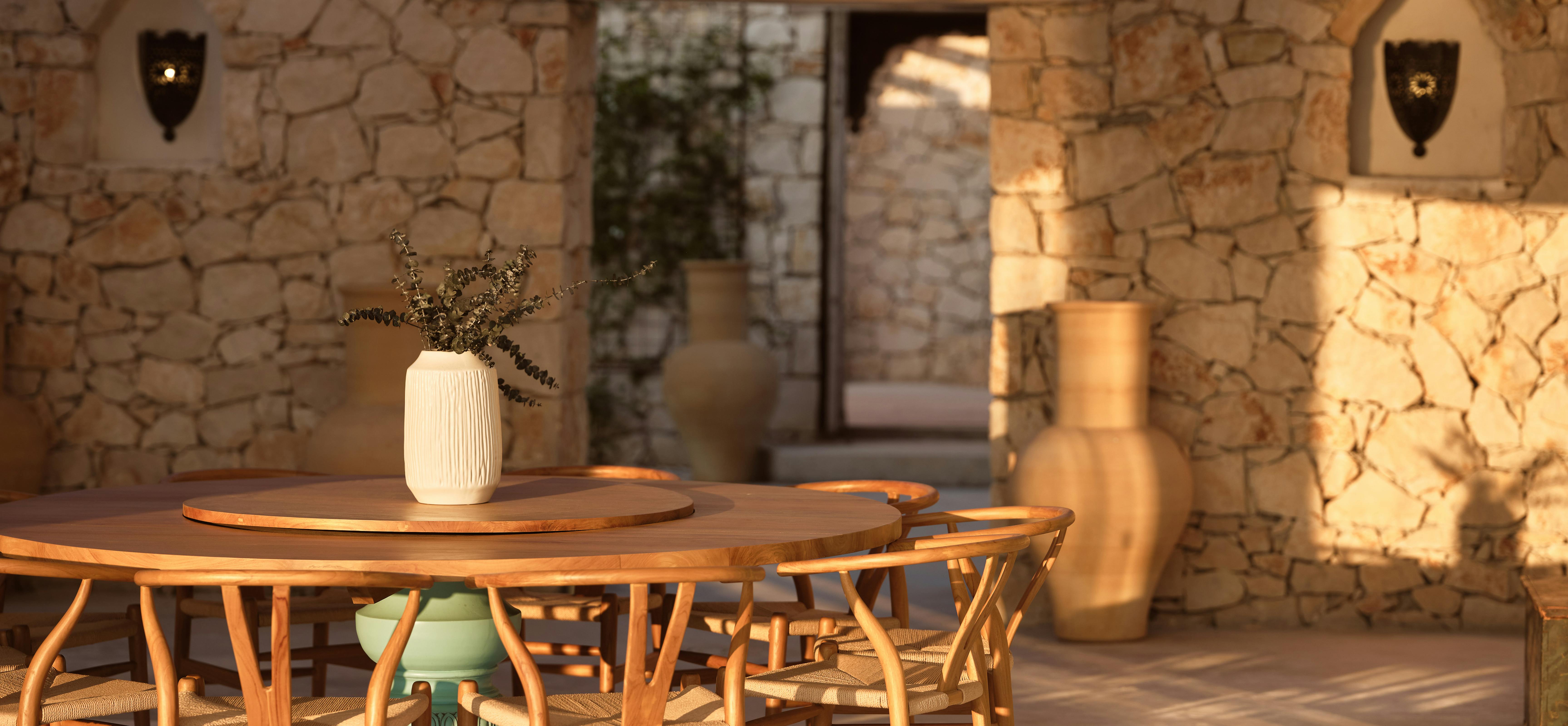 Warm evening light illuminates an outdoor dining area where a round wooden table surrounded by bent wishbone chairs sits beneath a large woven pendant lamp, with ancient stone walls framing views to an adjacent living space and garden beyond.