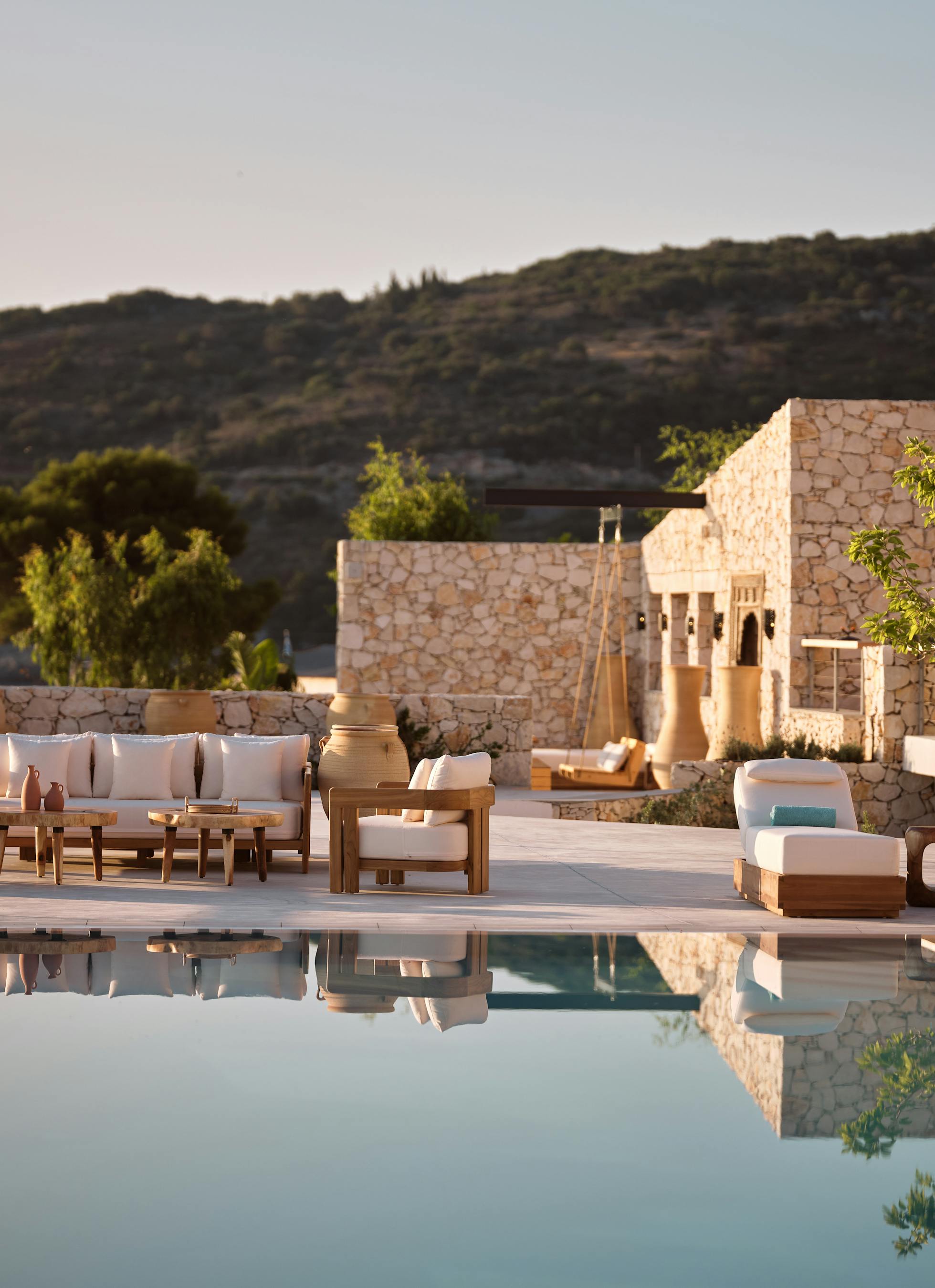 The glassy surface of an infinity pool creates perfect mirror images of the modern stone villa complex featuring clean geometric volumes, multiple terraced levels with white-cushioned loungers, large terracotta planters, and lush Mediterranean vegetation against distant hills.