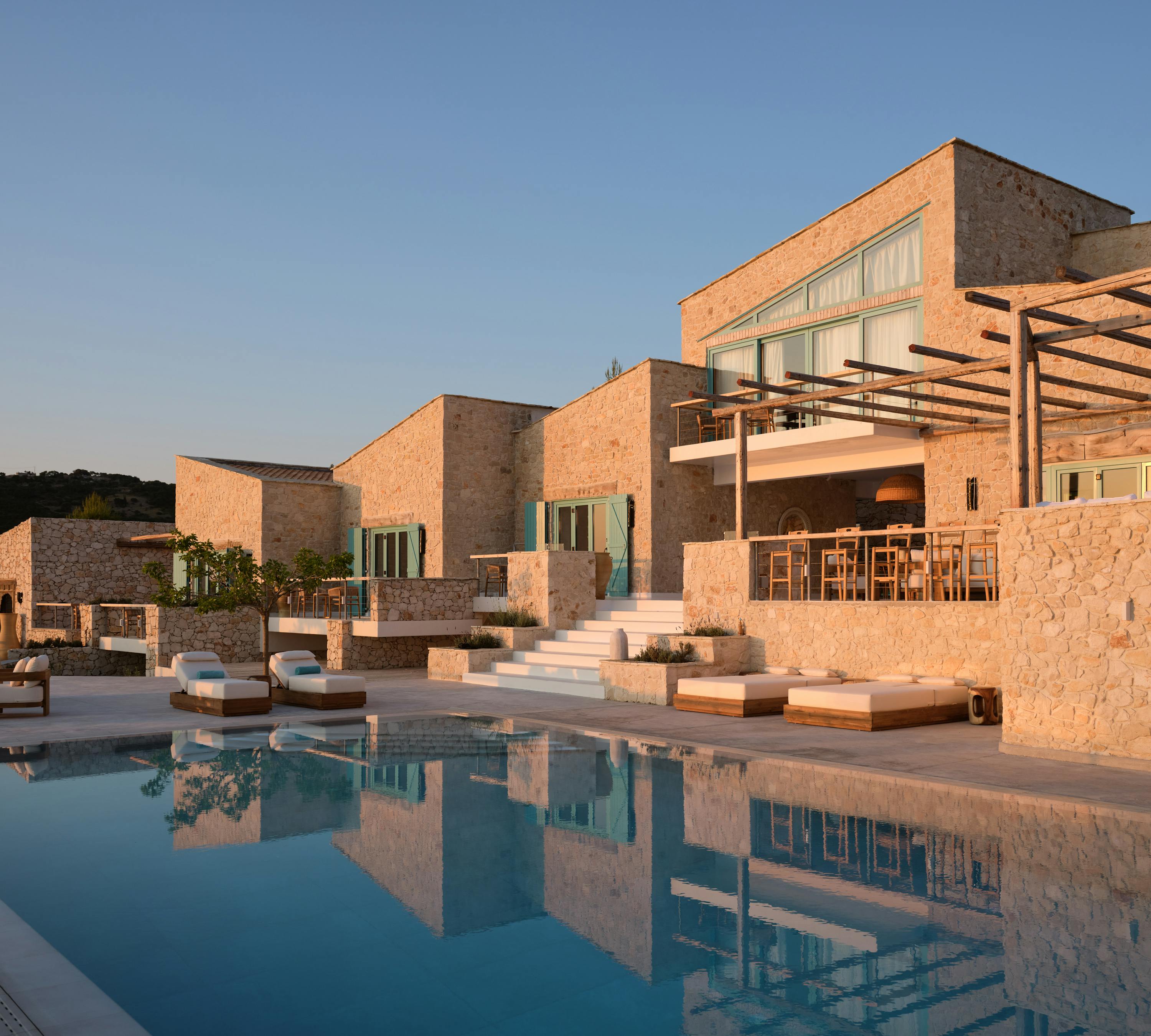 The infinity pool's turquoise water creates stunning reflections of the multi-story limestone villa complex bathed in golden evening light, showcasing the terraced architecture with mint-green shuttered windows, wooden pergolas, white stone steps, and scattered teak loungers.
