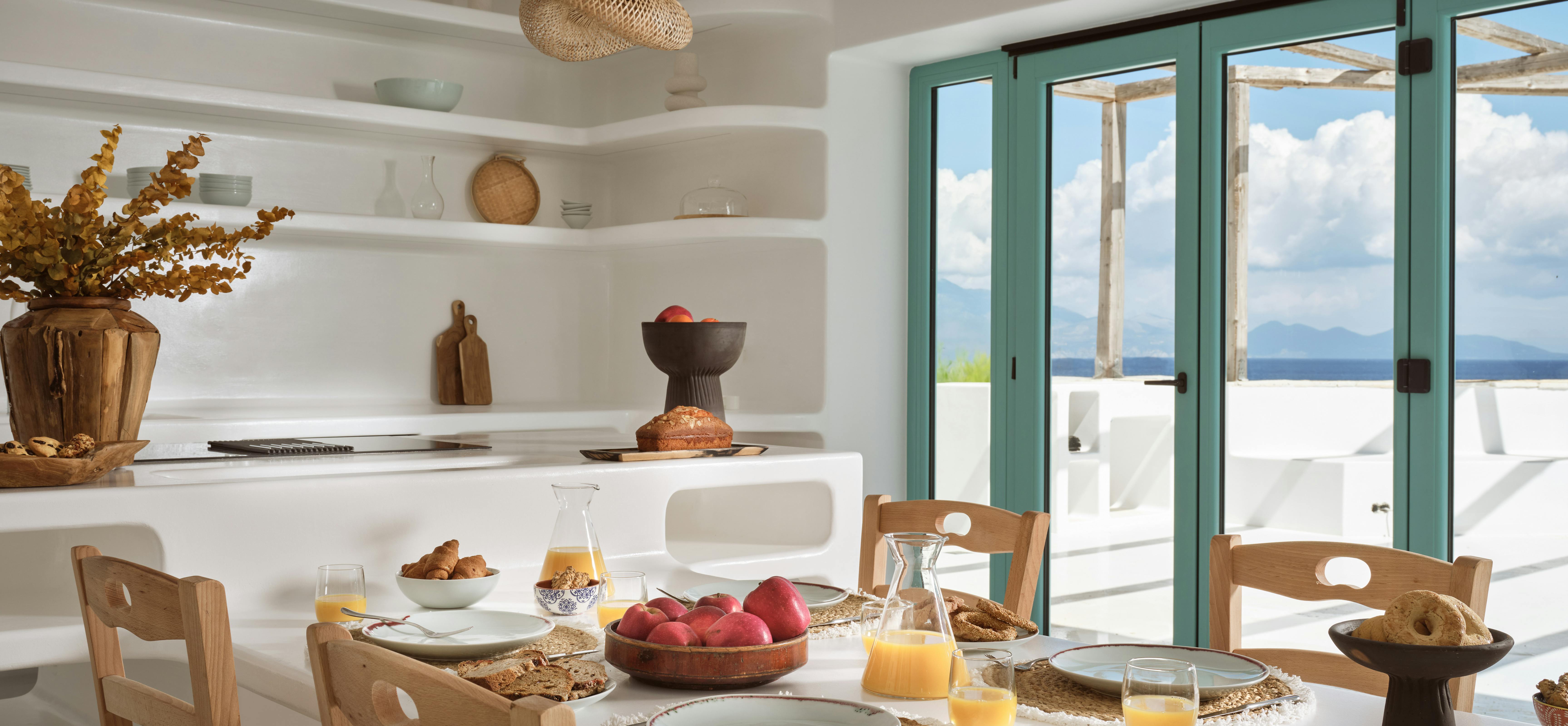 Dining area with white curved built-in bench, wooden chairs, and turquoise doors opening to sea terrace.