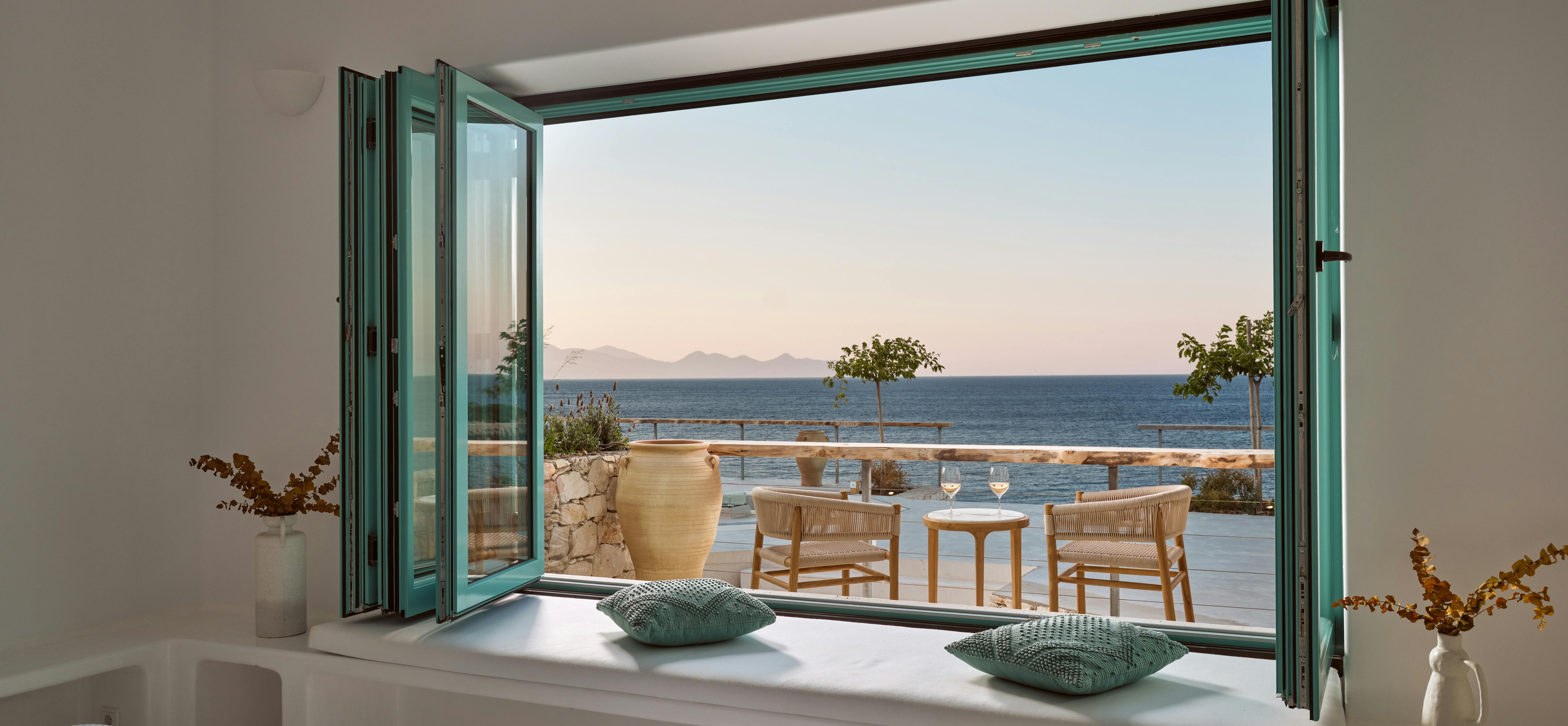 Bedroom with turquoise folding windows opening to furnished terrace overlooking the sea and distant islands.