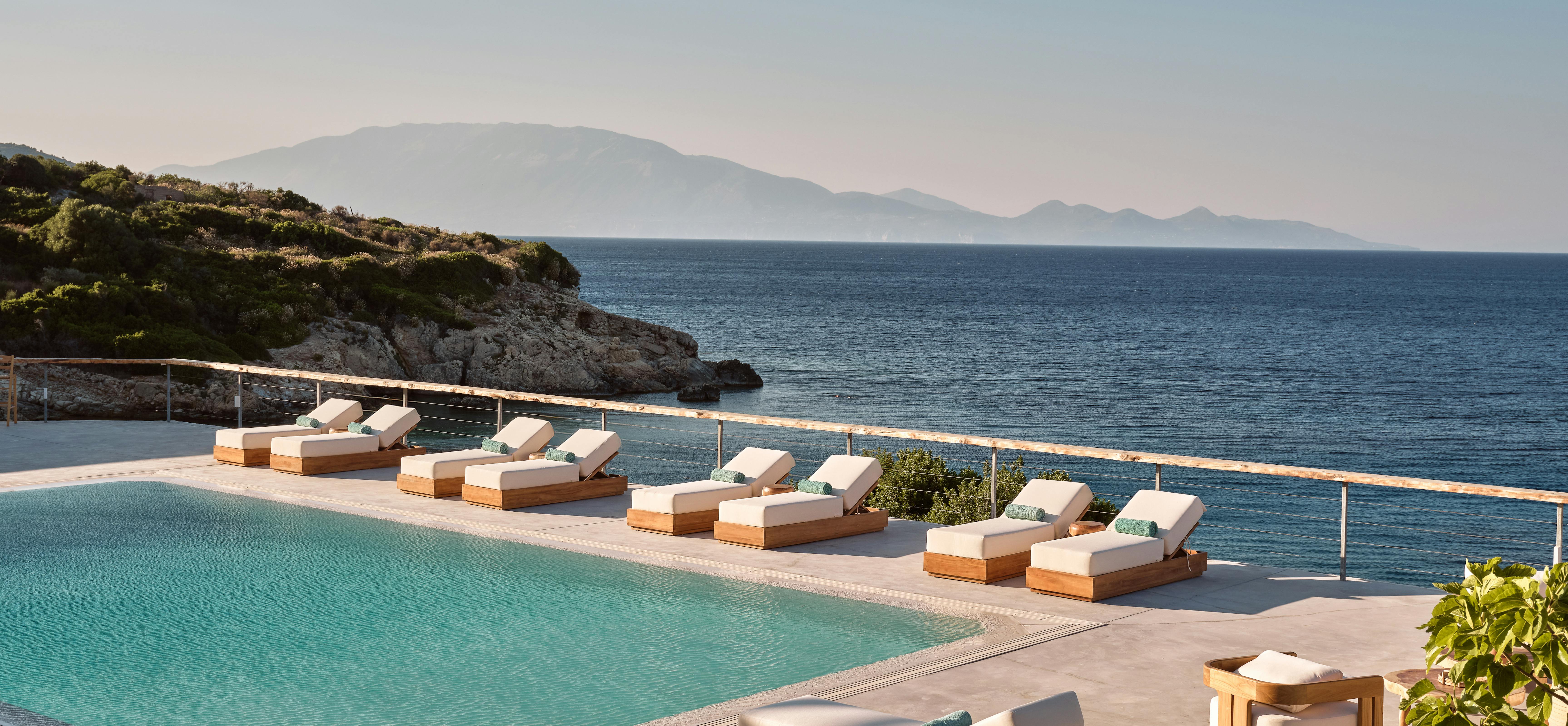 Infinity pool deck with wooden loungers overlooking calm blue sea and mountain silhouettes at sunset.