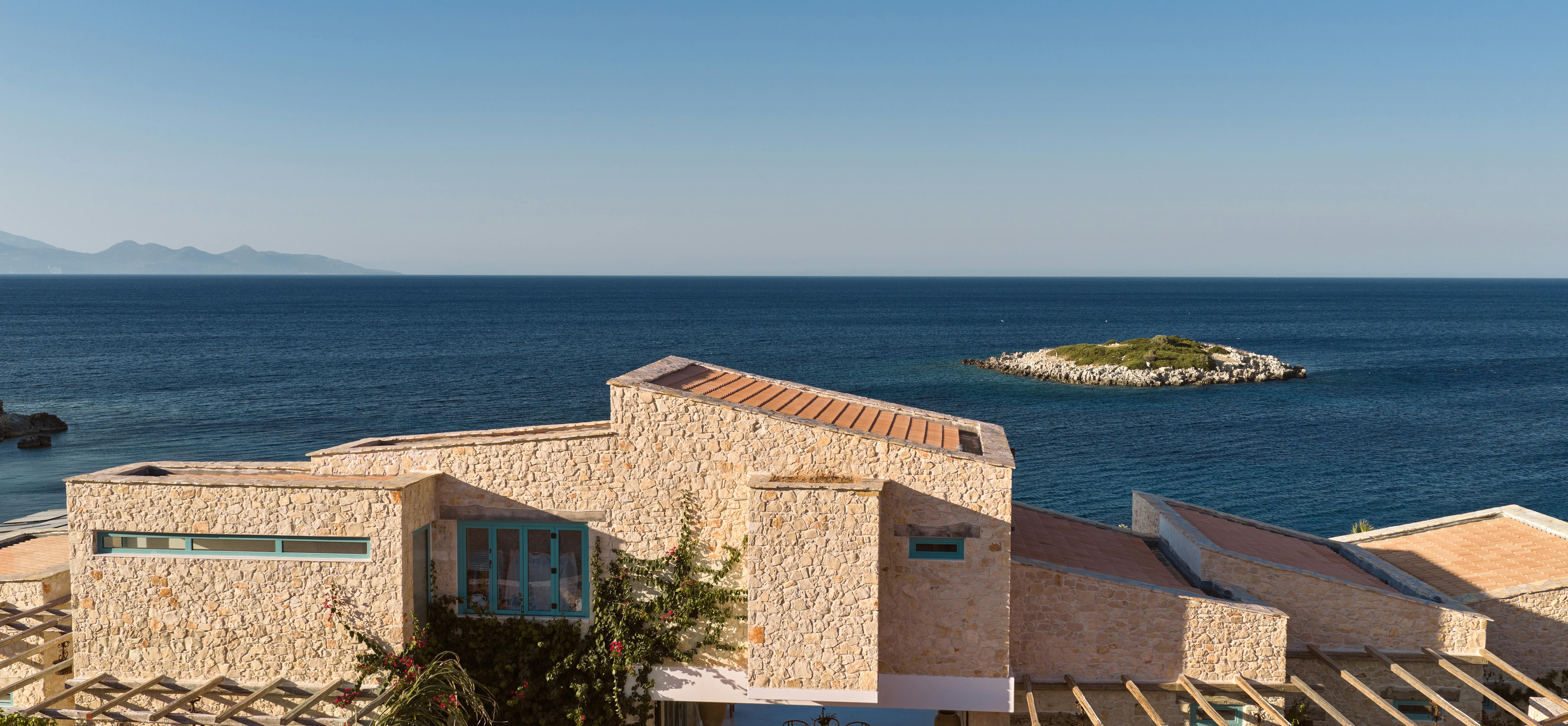 Exterior view of stone villa with turquoise windows perched on rocky Mediterranean coastline.