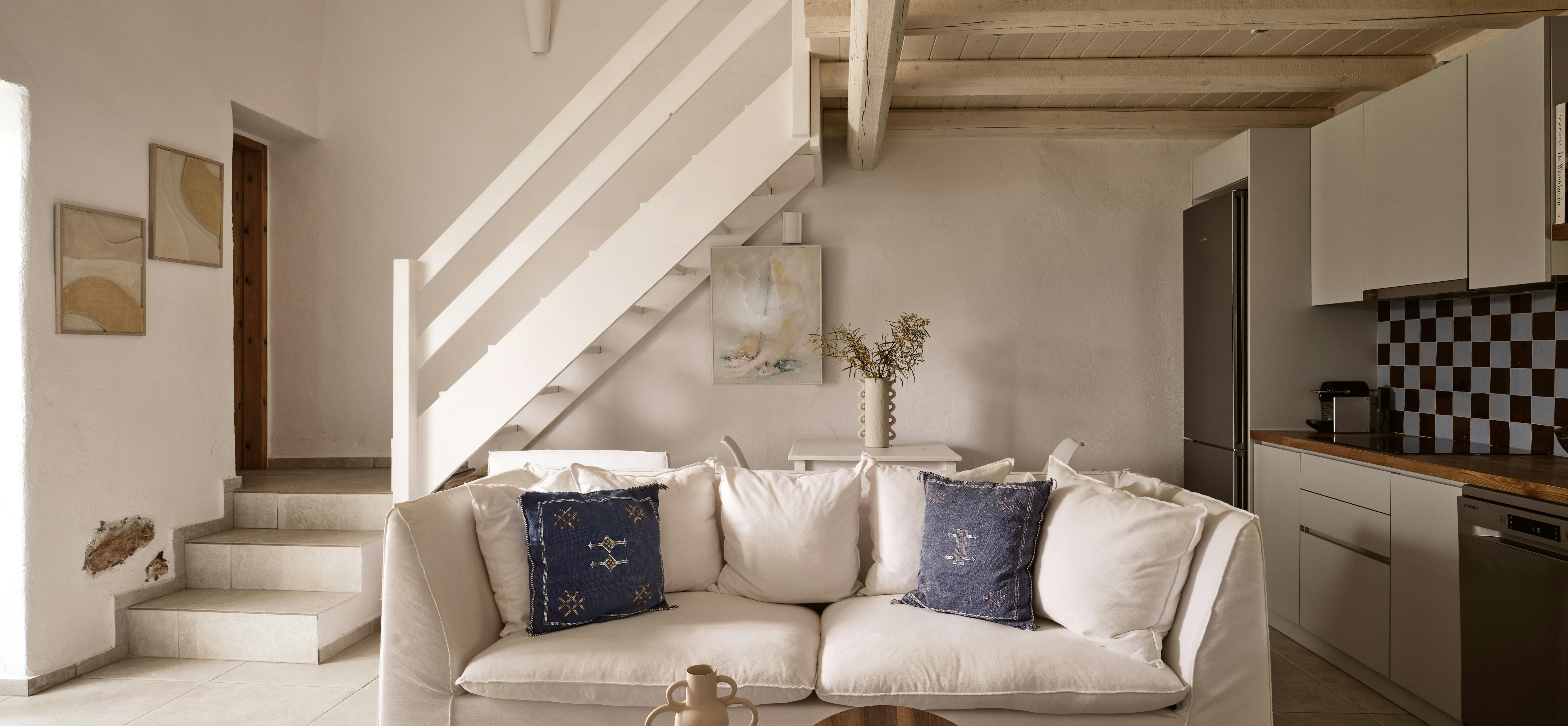 A bright Mediterranean living room with white walls, exposed wooden beam ceiling, cream sofa with blue pillows, and a staircase leading to an upper level, with a modern kitchen visible in the background.