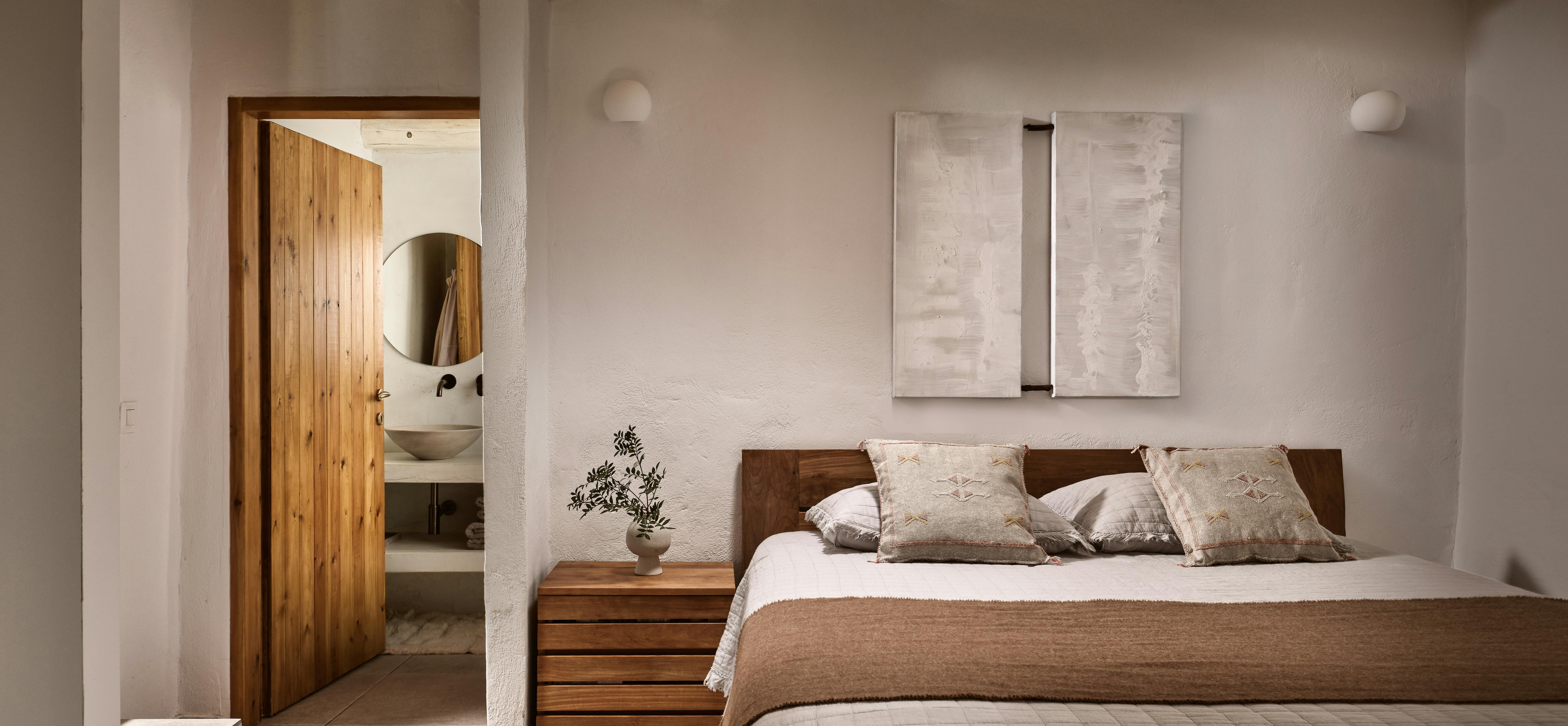 A minimalist bedroom featuring a wooden platform bed with brown and cream bedding, exposed wooden ceiling beams, white walls, abstract artwork, and a glimpse of an ensuite bathroom through an open doorway.