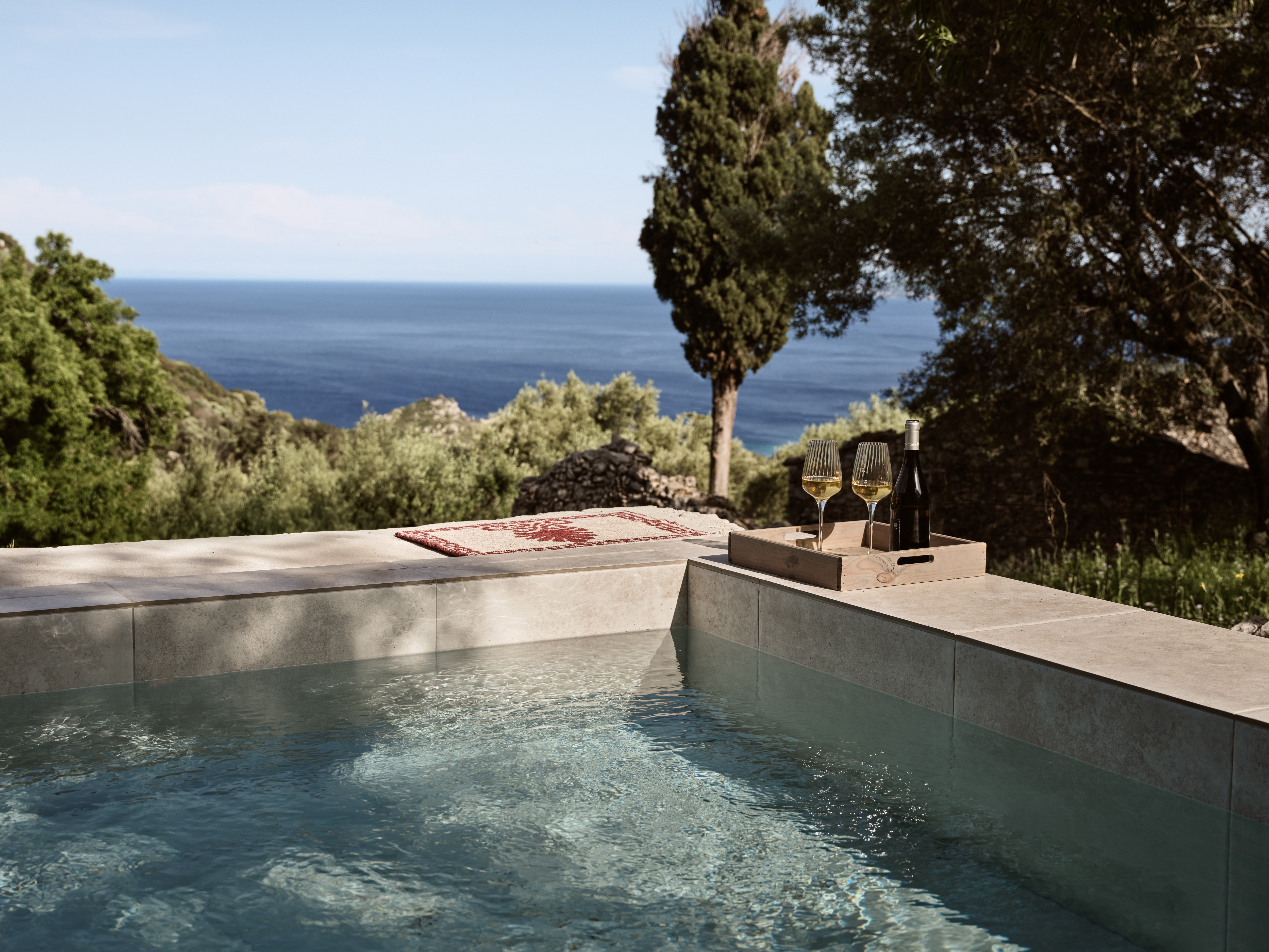 An infinity pool edge with concrete surround displaying wine glasses and a bottle on a tray, overlooking the deep blue Mediterranean Sea with cypress trees framing the view.