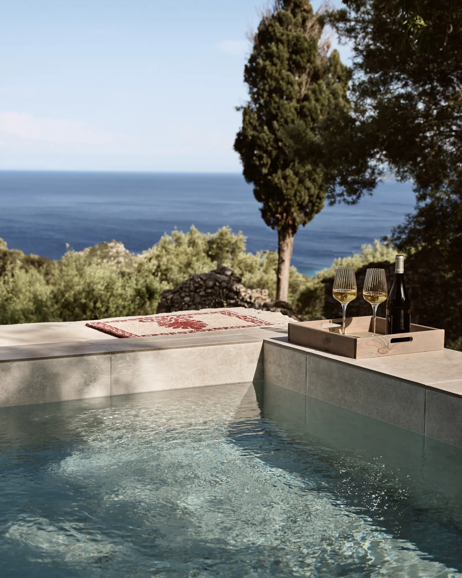 An infinity pool edge with concrete surround displaying wine glasses and a bottle on a tray, overlooking the deep blue Mediterranean Sea with cypress trees framing the view.