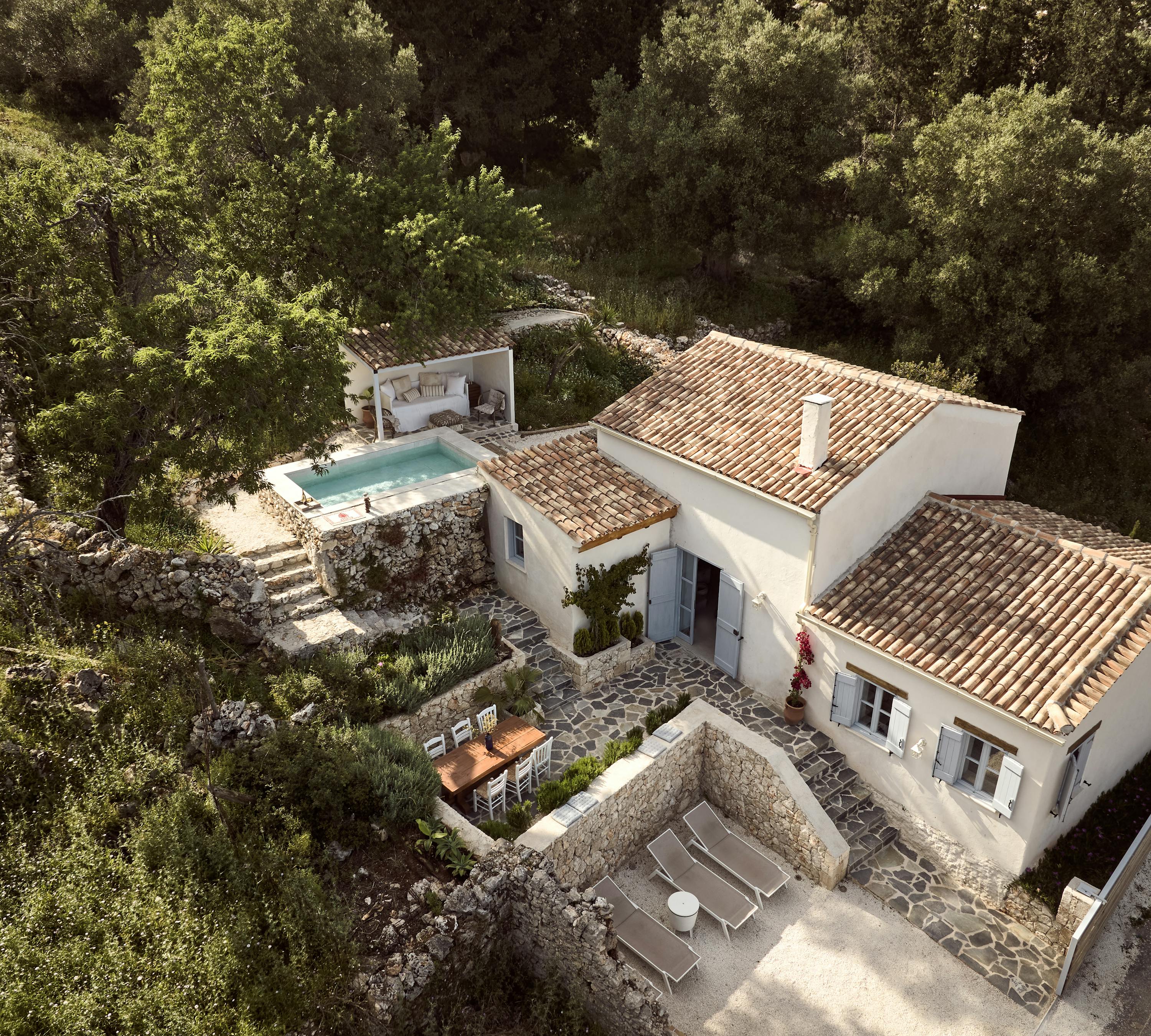 An aerial view of a white Mediterranean villa with terracotta tile roofing nestled in lush greenery, featuring a pool with a covered lounge area and stone terraced outdoor spaces.