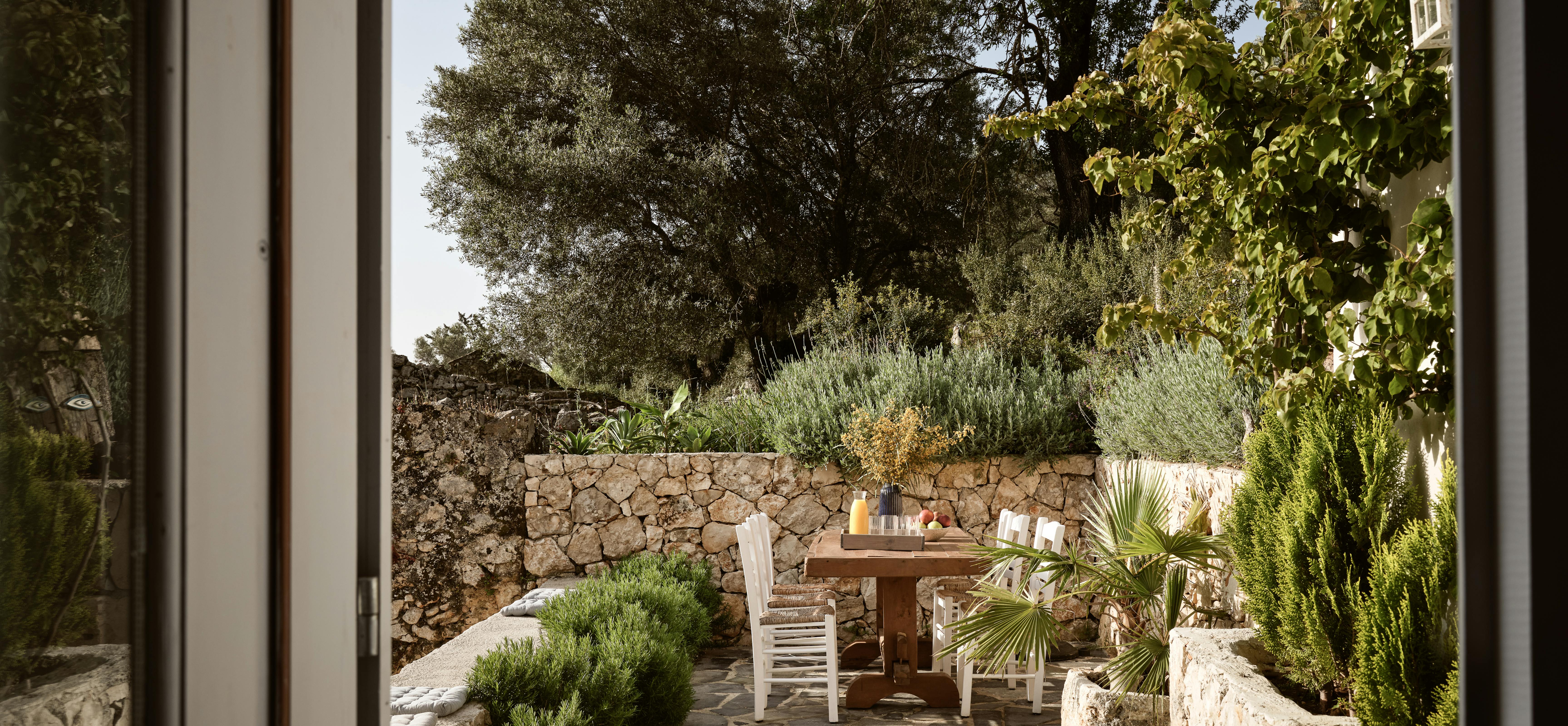 A stone-paved outdoor dining terrace surrounded by dry-stacked stone walls and Mediterranean vegetation, with a wooden table and white chairs set for al fresco dining.
