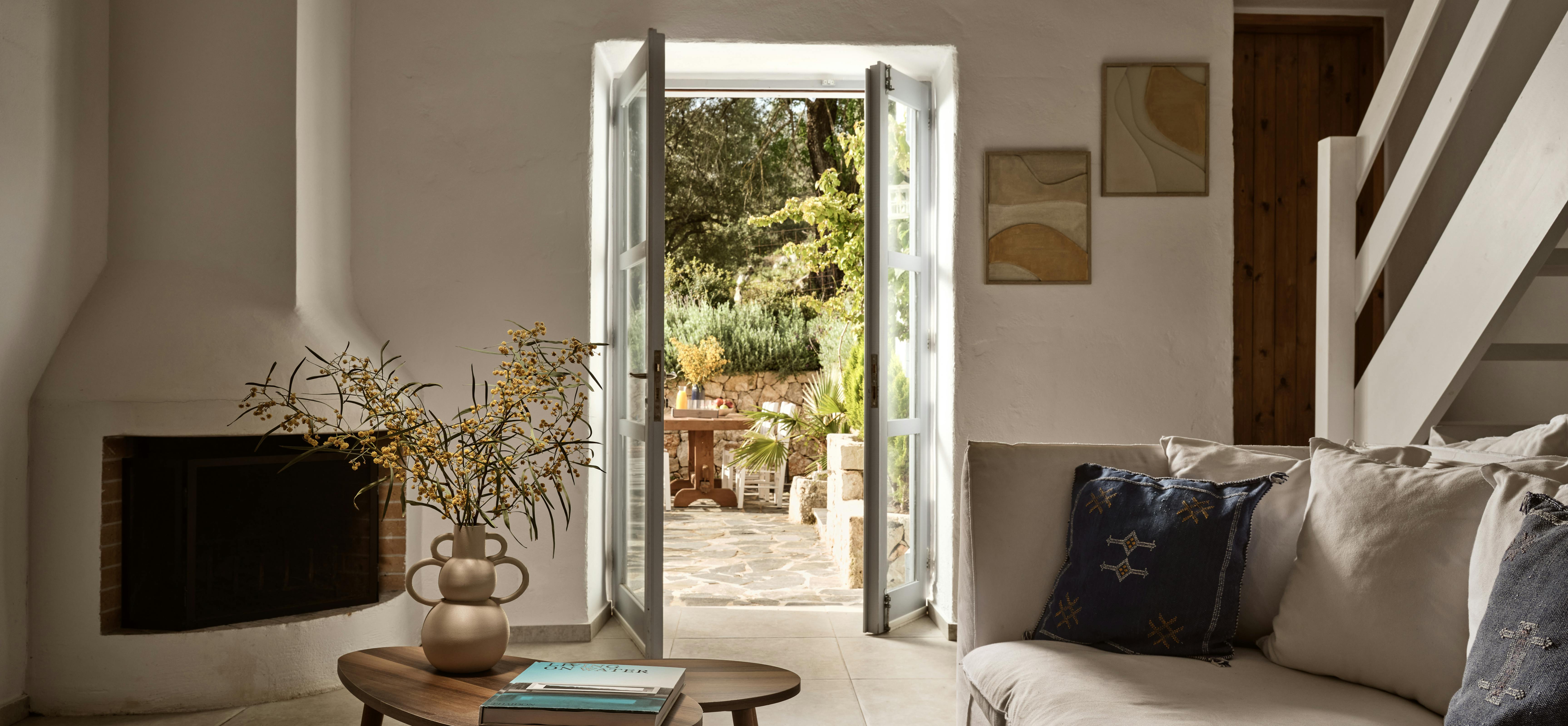 A serene living room with white walls, a modern fireplace, cream sofa with decorative pillows, and French doors opening onto a sunny terrace with outdoor dining furniture visible beyond.