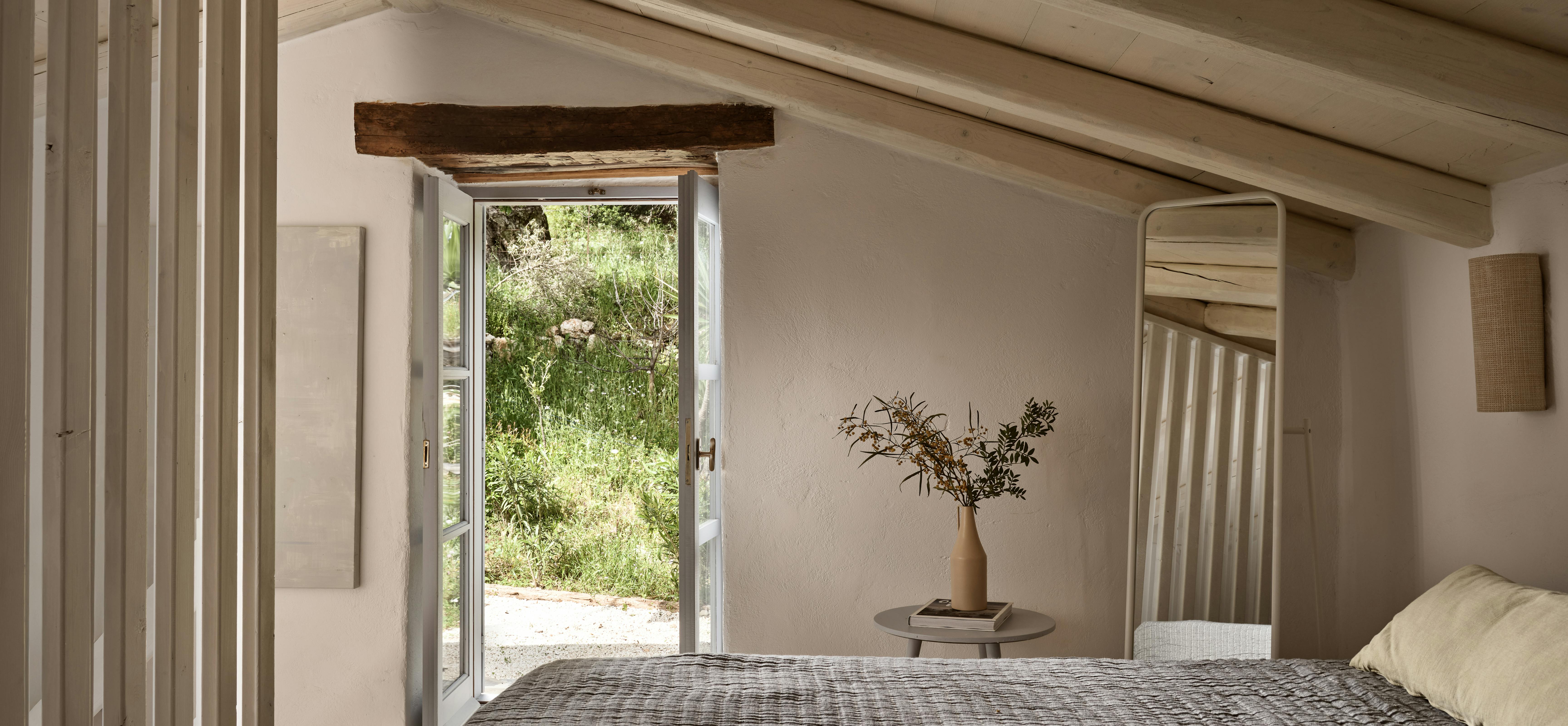 A bedroom with vaulted wooden plank ceiling, white walls, an open doorway framing a view of wild garden beyond, and a bed with grey quilted bedding.