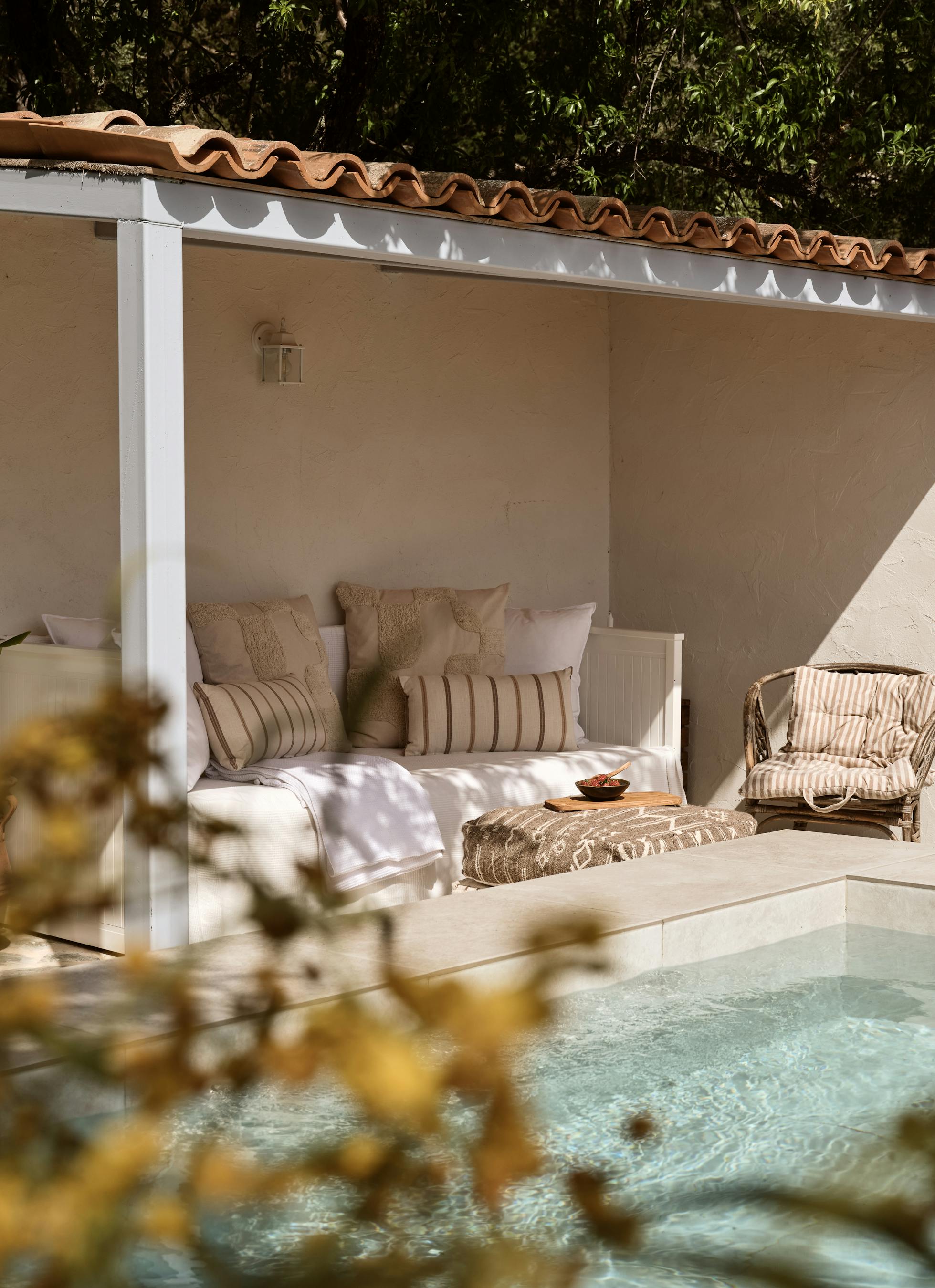 A covered outdoor daybed with white cushions and striped pillows beside a pool, set within a white-walled pavilion with terracotta tile roof surrounded by Mediterranean gardens.