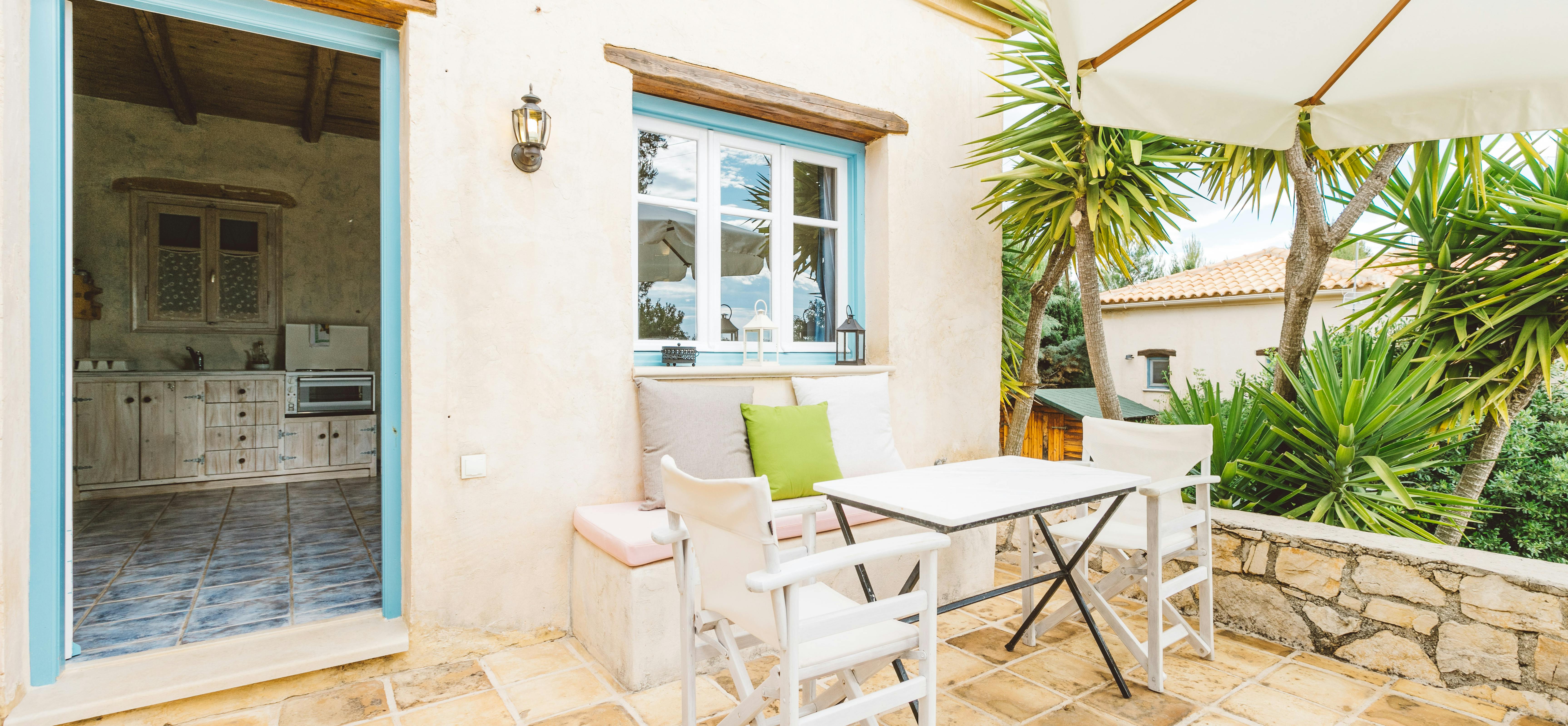 A sunny Mediterranean terrace with white walls, turquoise-trimmed windows and doors, white folding chairs around a small dining table, palm trees, and stone paving.
