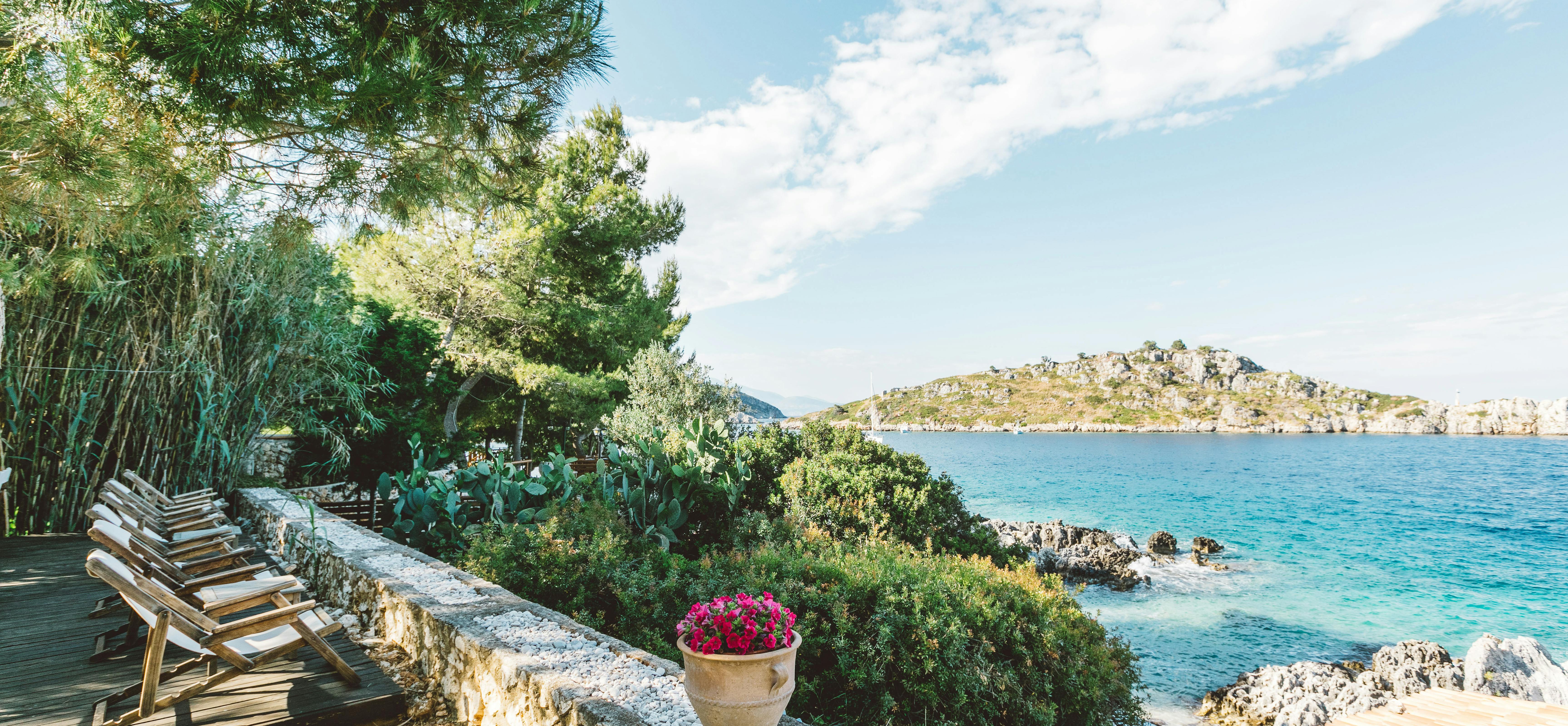 A coastal terrace with wooden sun loungers overlooking turquoise Mediterranean waters, a rocky island, lush vegetation, and a terracotta-roofed structure visible on the shoreline.