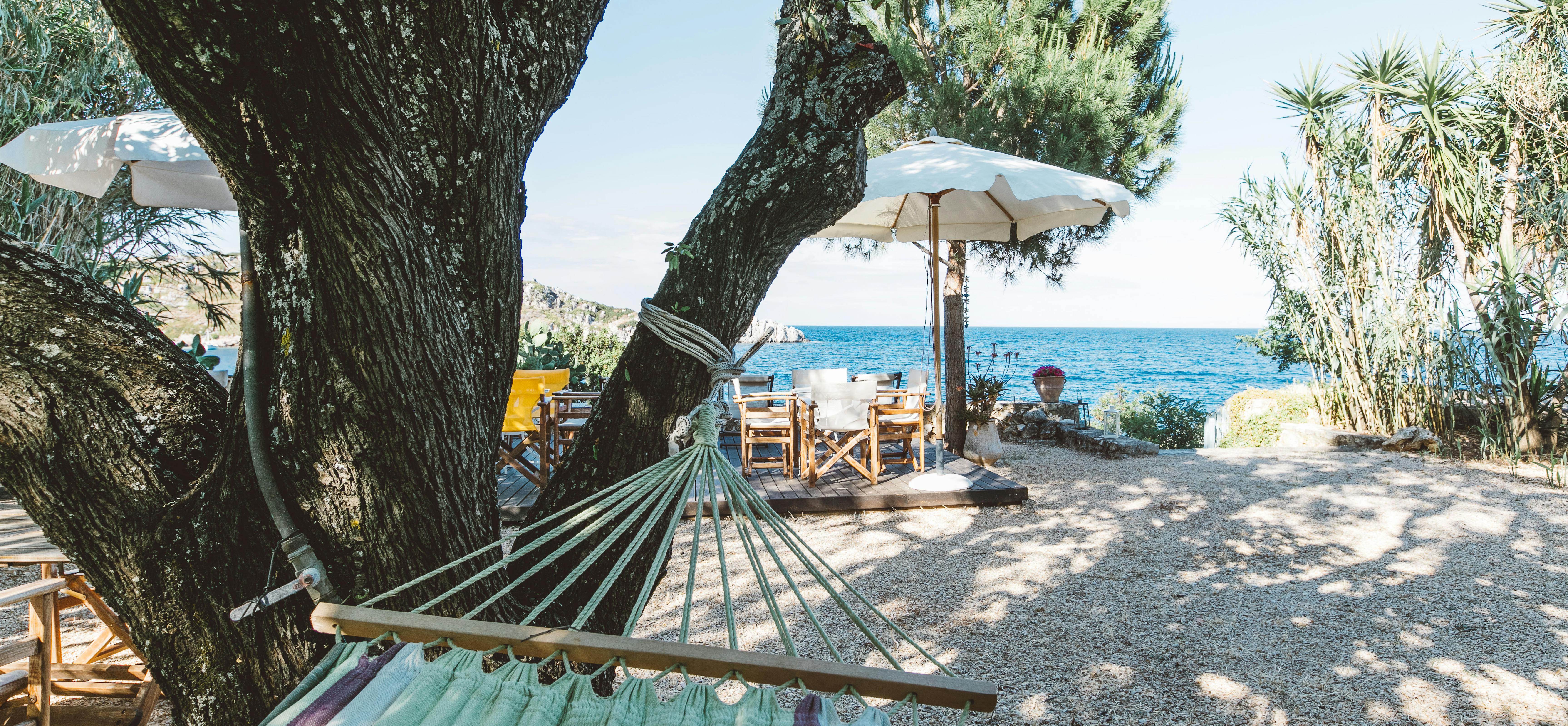 A turquoise and navy striped hammock hangs from a large olive tree in a sun-dappled beachfront garden, with canvas director's chairs under white umbrellas and the Mediterranean Sea visible beyond.