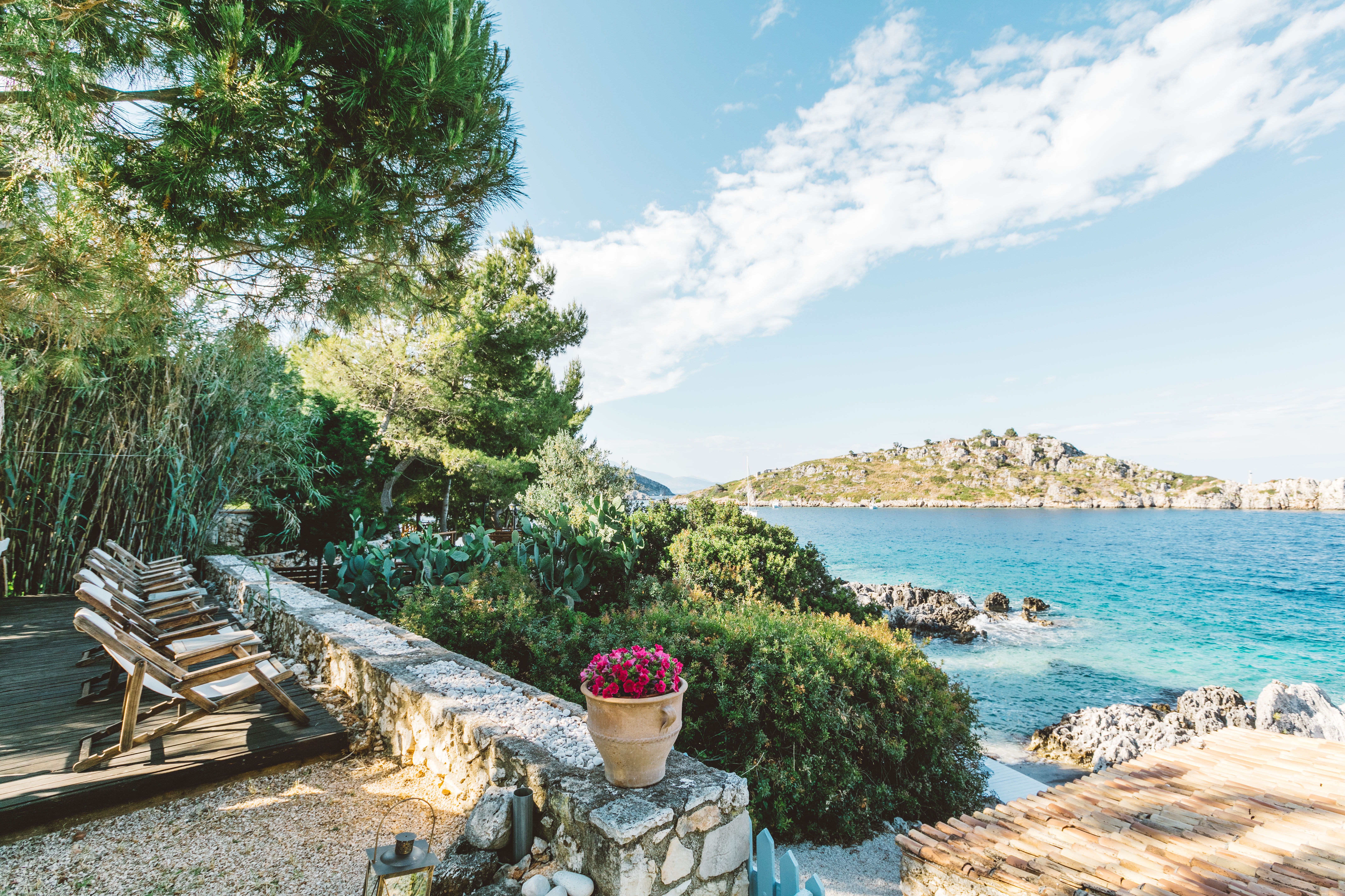 A coastal terrace with wooden sun loungers lined along a low stone wall, a terracotta pot with pink flowers, lush vegetation, and views of turquoise Mediterranean waters and a rocky island.