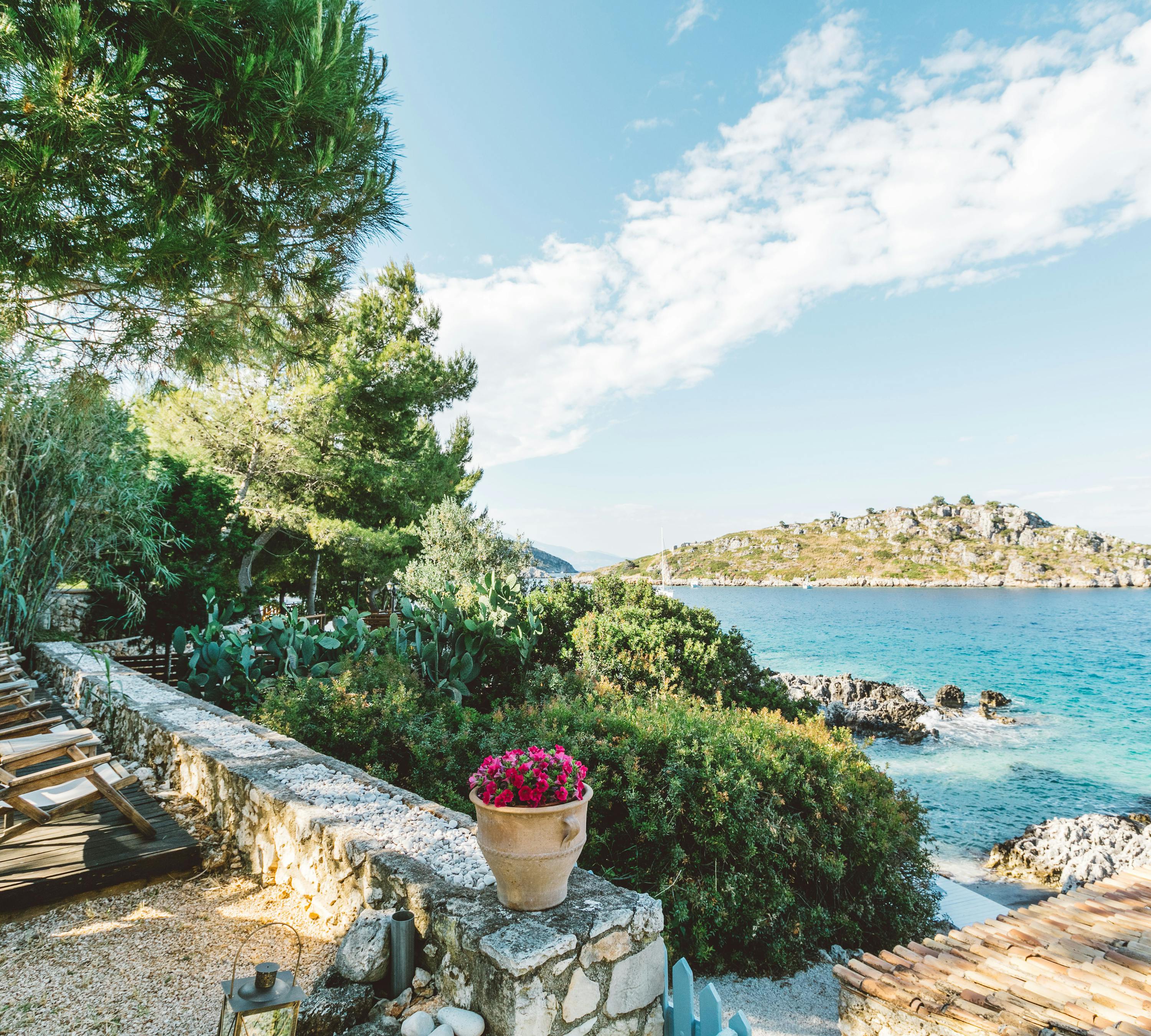 A coastal terrace with wooden sun loungers lined along a low stone wall, a terracotta pot with pink flowers, lush vegetation, and views of turquoise Mediterranean waters and a rocky island.