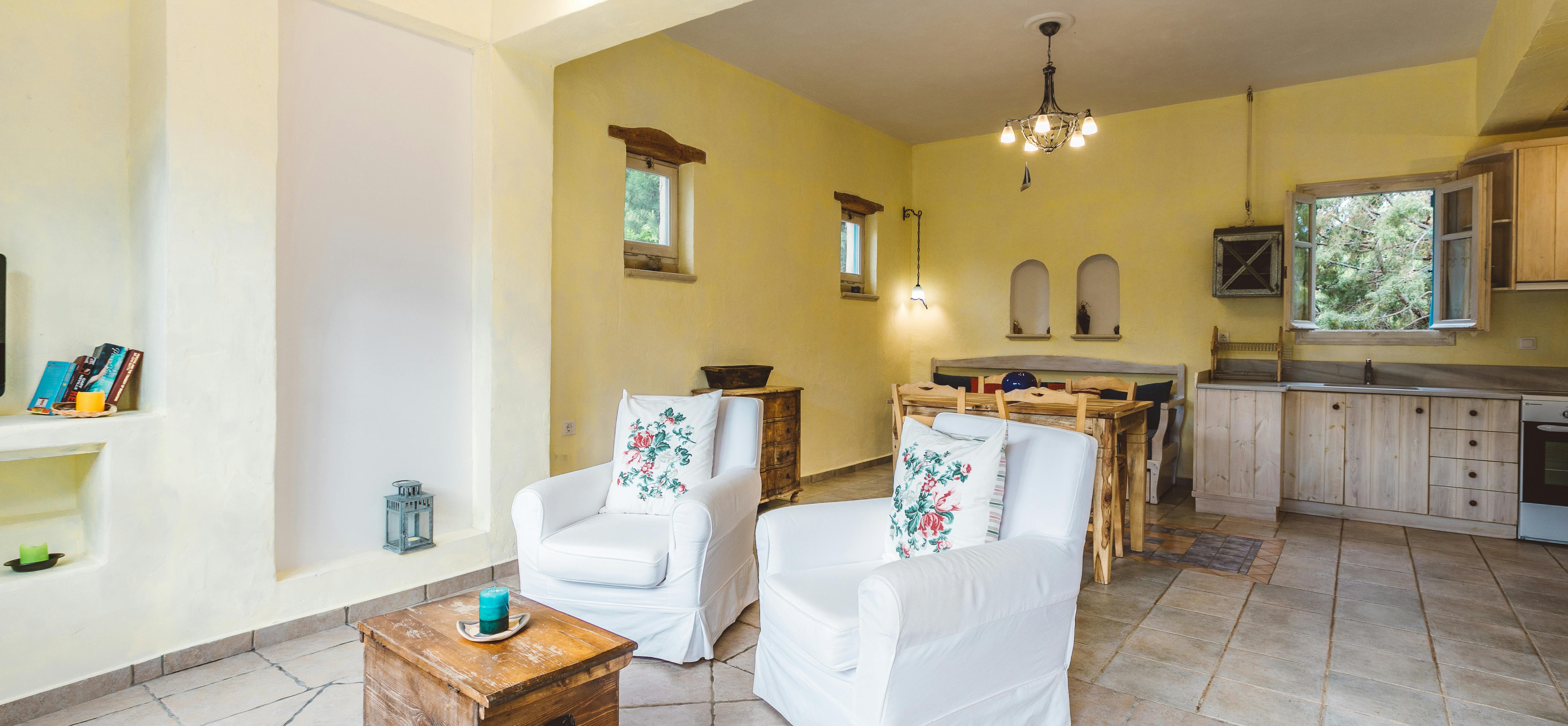 An open-plan Mediterranean living space with yellow and white walls, stone tile floor, two white slipcovered chairs with floral pillows, wooden dining table, built-in niches, and kitchen visible in background.