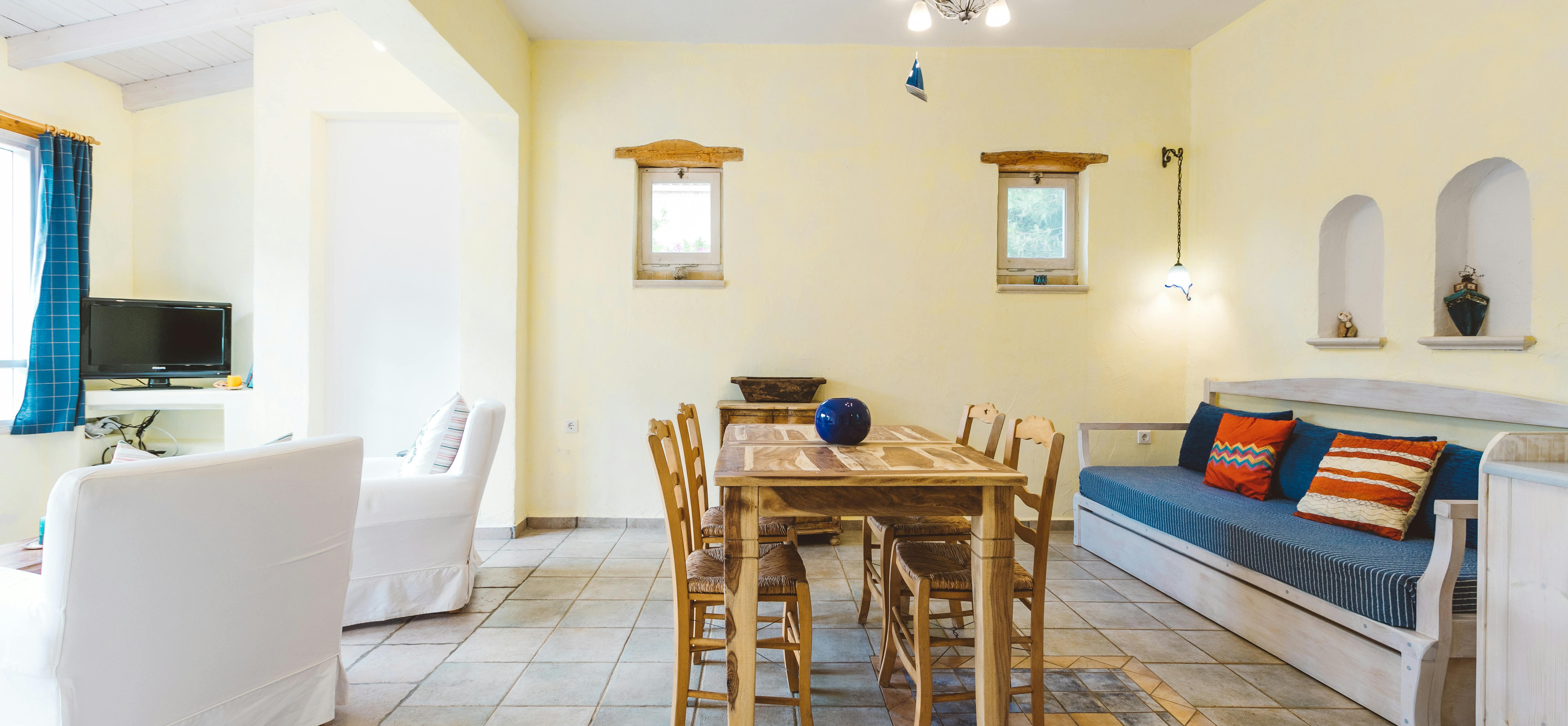 A spacious open-plan Mediterranean interior with yellow walls, white slipcovered chairs, wooden dining table, platform daybed with blue and striped pillows, arched niches, and multicoloured tile floor.