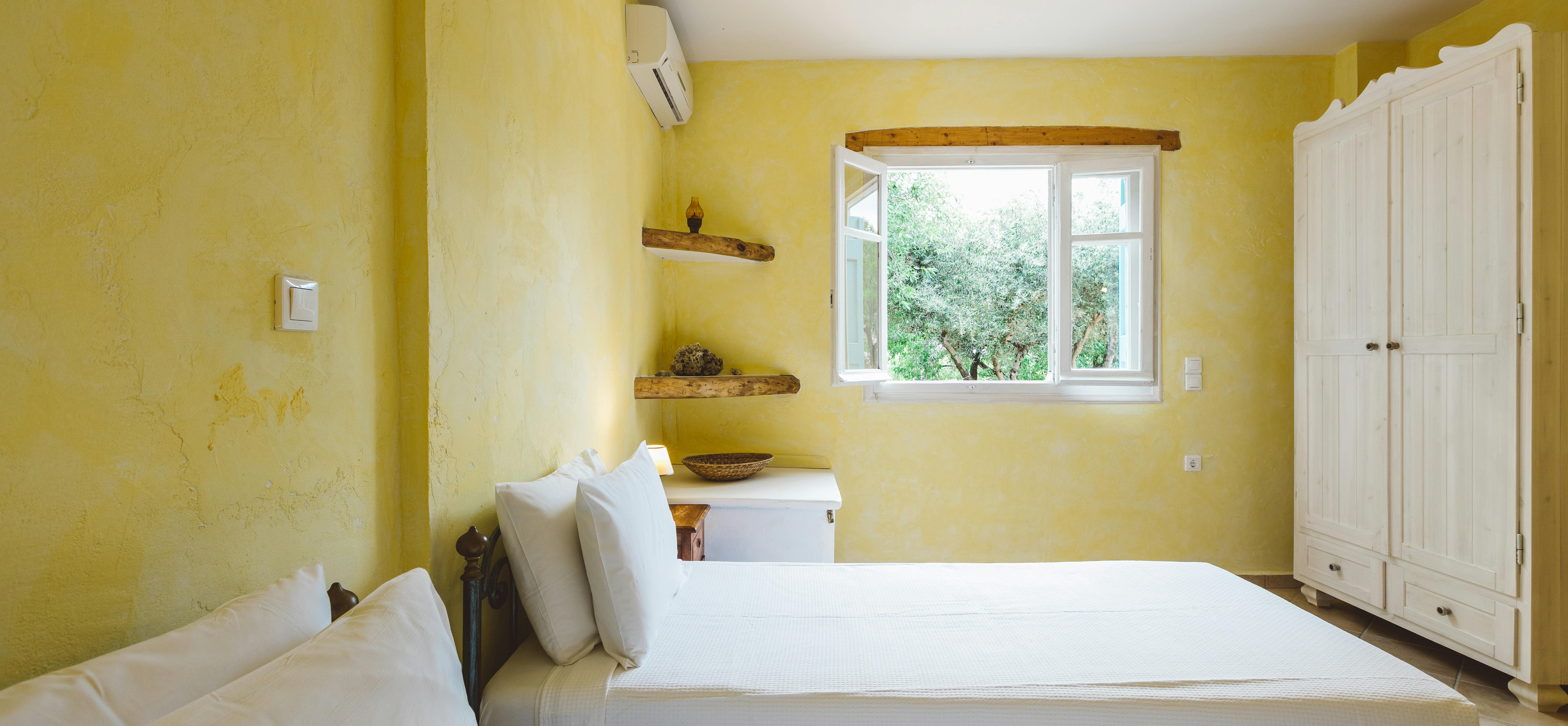 A cheerful yellow bedroom with twin white platform beds, wooden floating shelves, white wardrobe, window overlooking olive trees, air conditioning unit, and pale tile floor.
