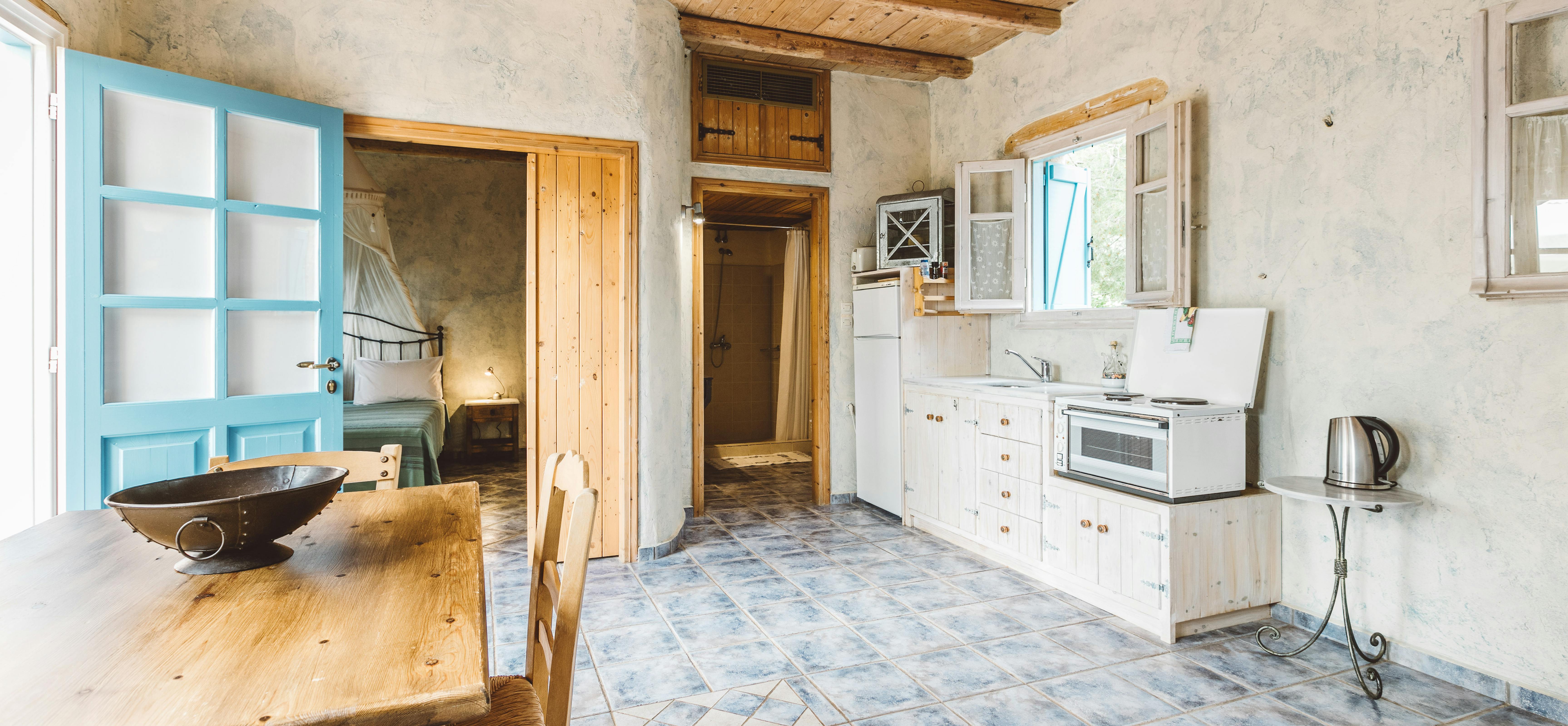 A rustic kitchen-dining area with distressed plaster walls, exposed wooden ceiling, turquoise door, wooden dining table, white kitchen cabinets, and blue patterned tile floor.