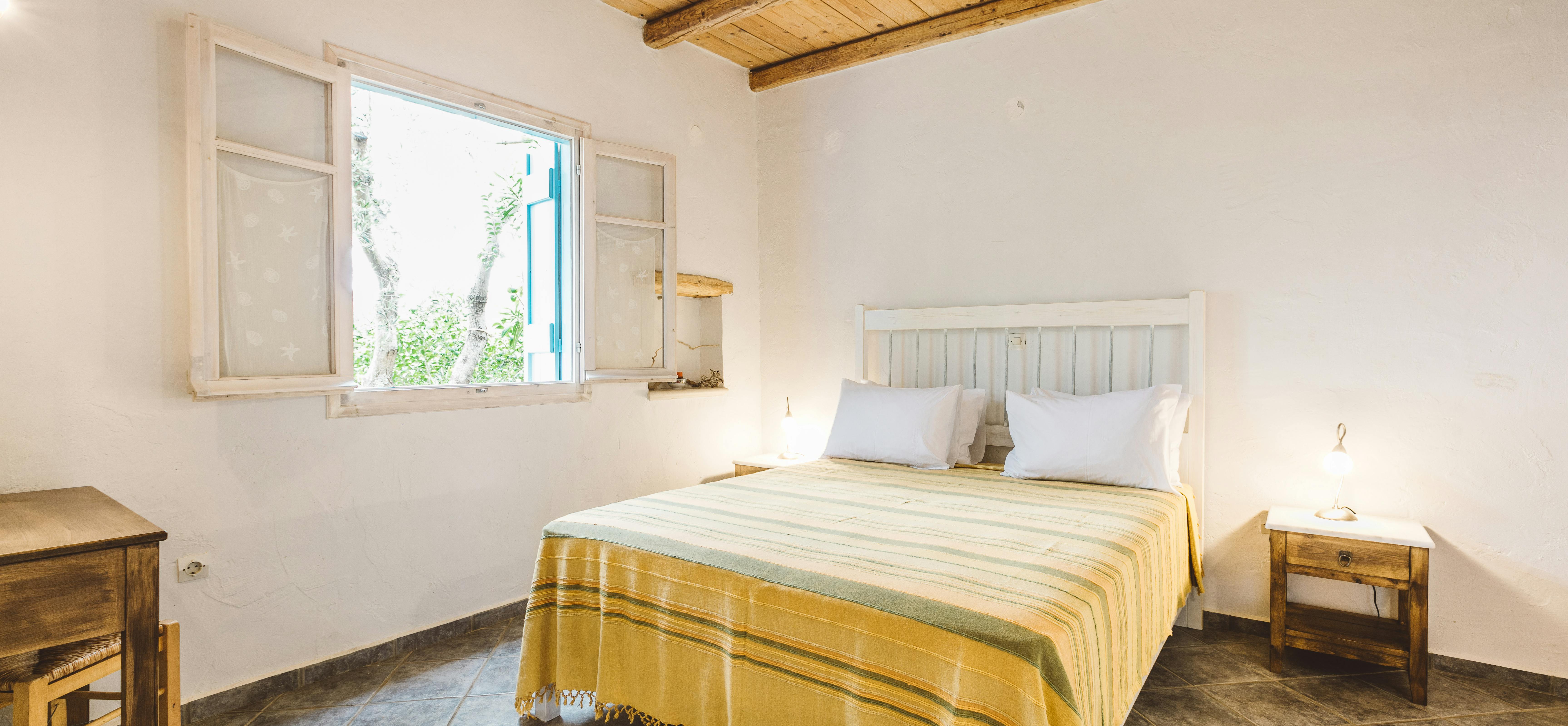 A minimalist white bedroom with exposed wooden ceiling beams, white painted wood headboard, bed with yellow and green striped throw, simple wooden furniture, and window overlooking olive trees.
