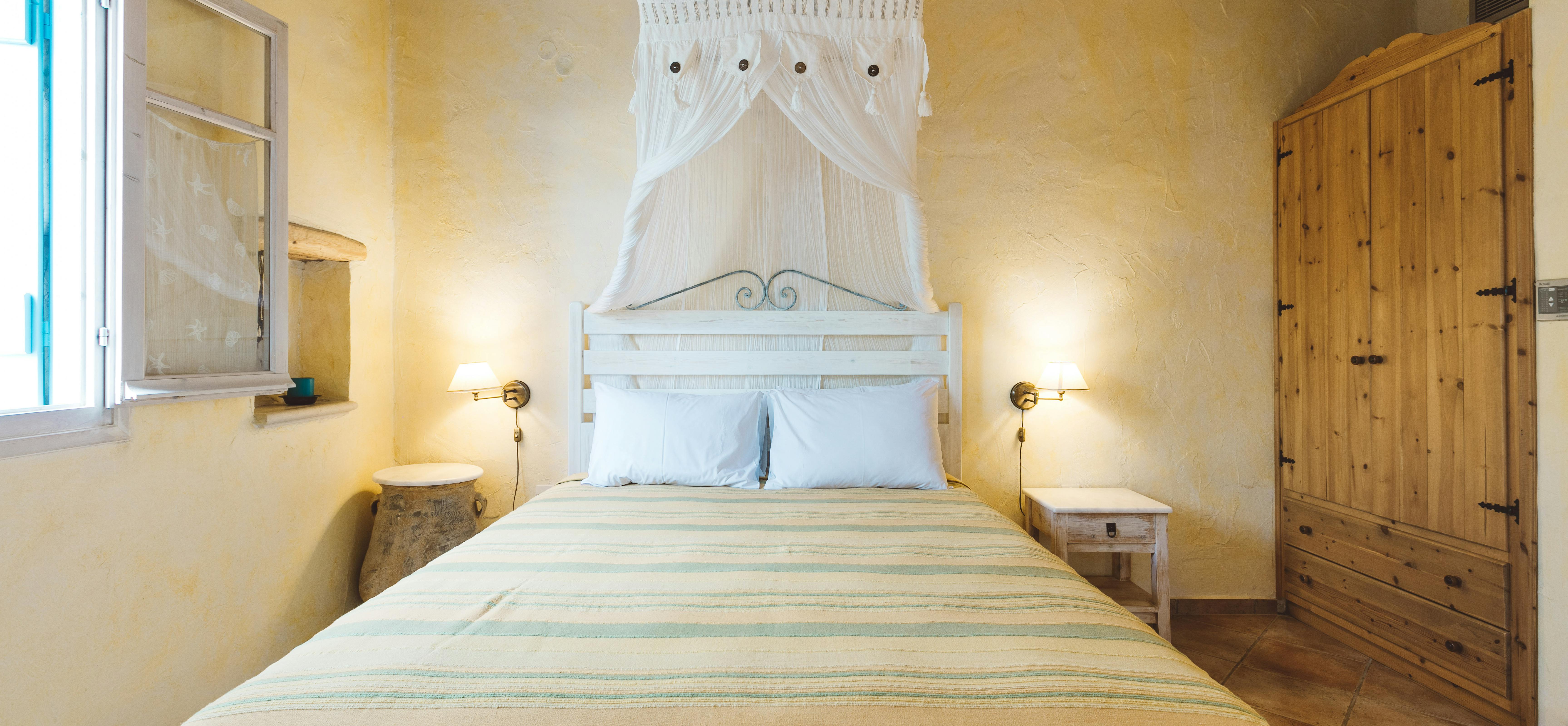 A sunny bedroom with pale yellow walls, exposed wooden ceiling, white painted wood headboard with white mosquito netting, bed with turquoise and yellow striped bedspread, wall sconces, and large wooden wardrobe.