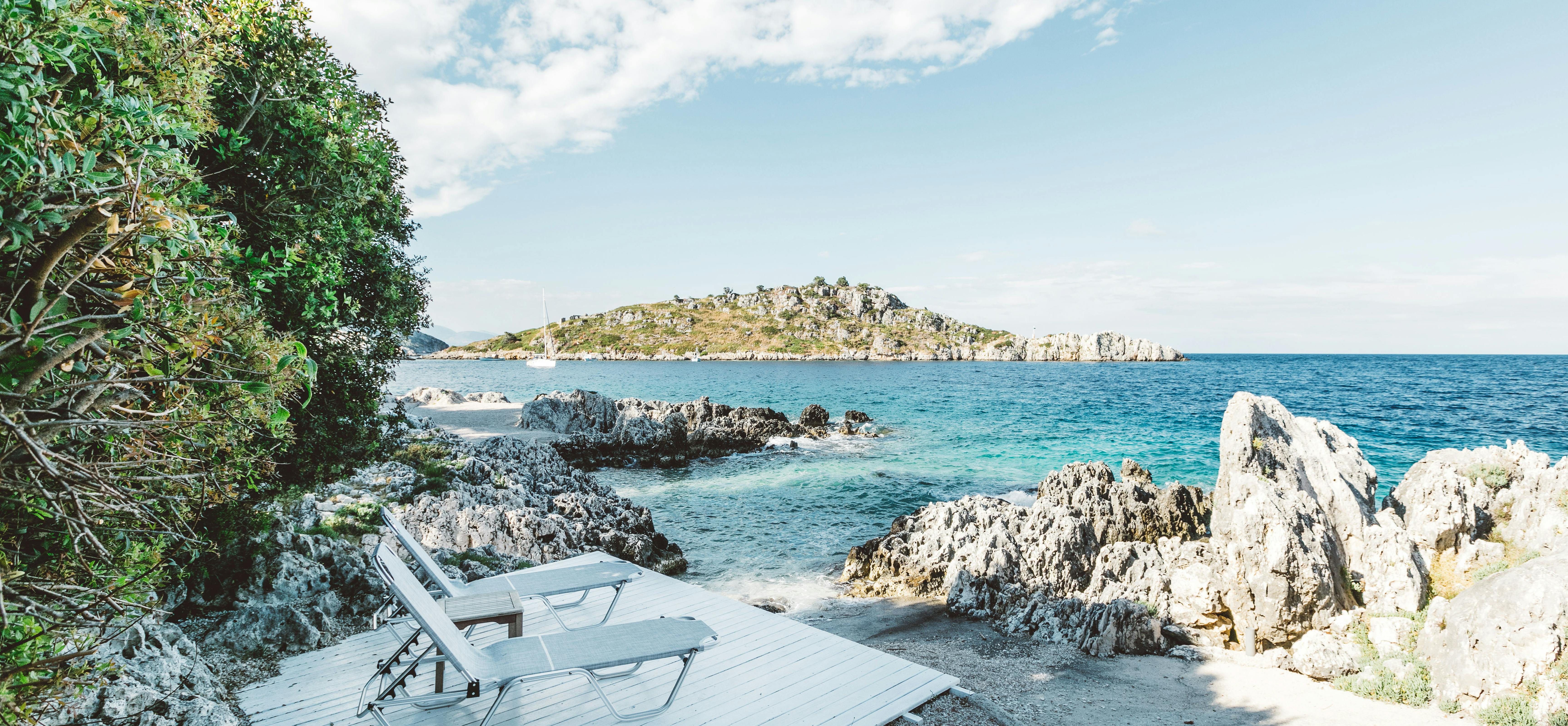 A secluded coastal deck with two white sun loungers on weathered wood overlooking turquoise waters, jagged white limestone rocks, a small beach, and a tree-covered island under blue skies.