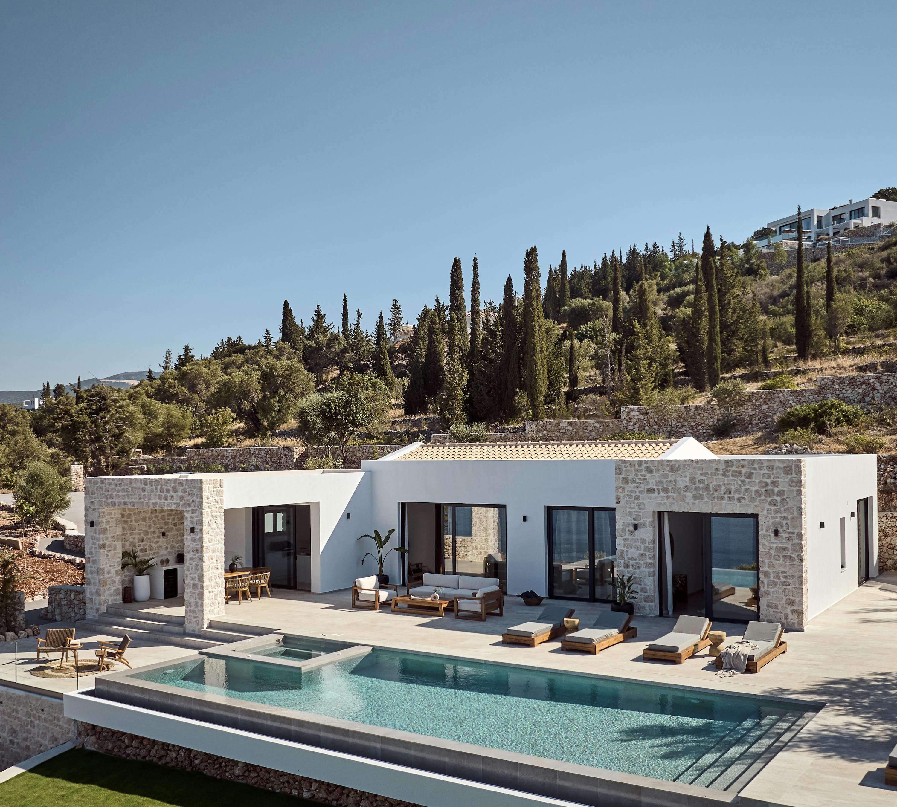 A modern hillside Mediterranean villa with white stucco and stone facades, featuring a large infinity pool surrounded by wooden sun loungers, terraced landscaping with cypress trees and olive groves climbing the slope behind.