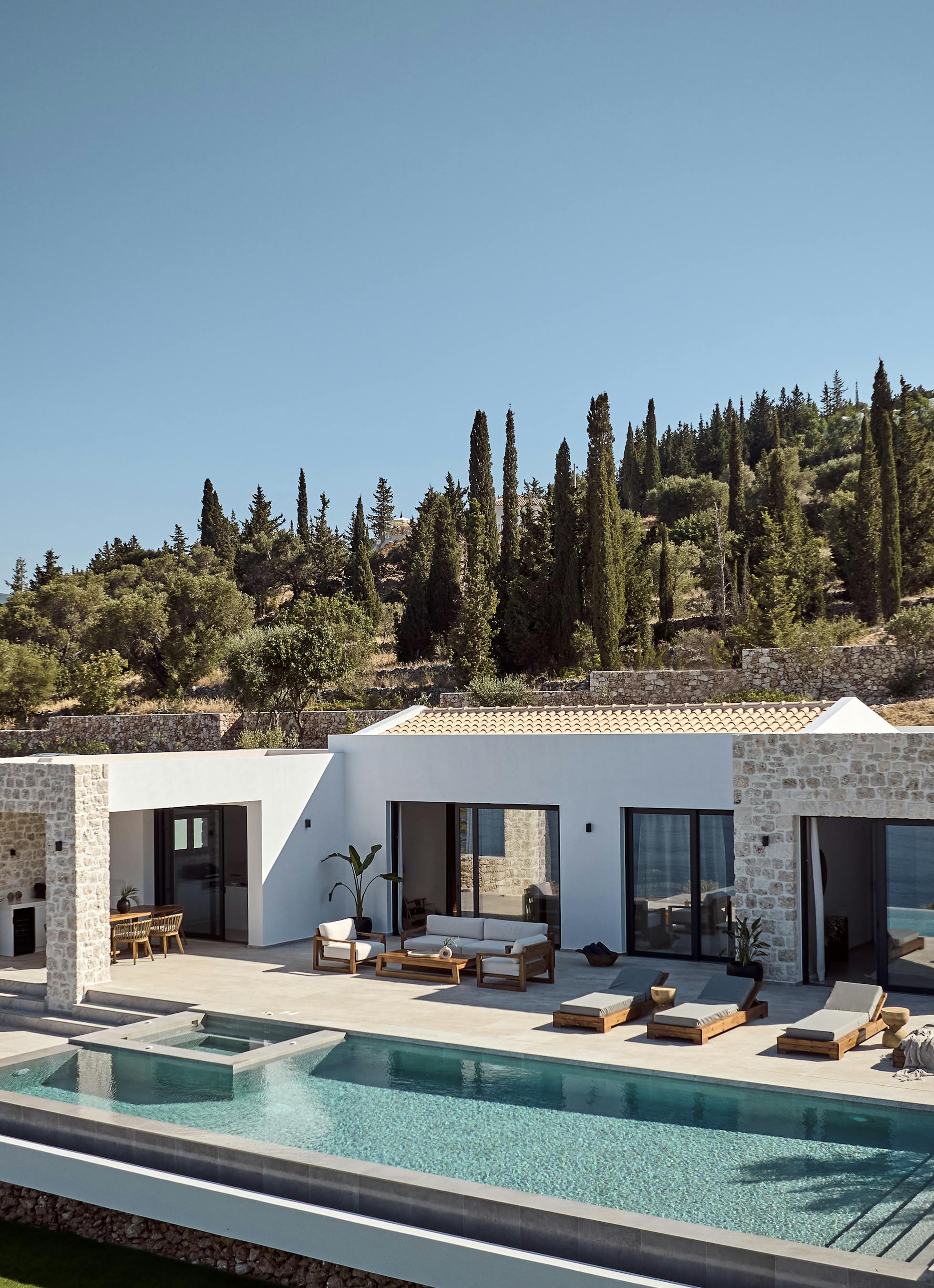 A modern hillside Mediterranean villa with white stucco and stone facades, featuring a large infinity pool surrounded by wooden sun loungers, terraced landscaping with cypress trees and olive groves climbing the slope behind.