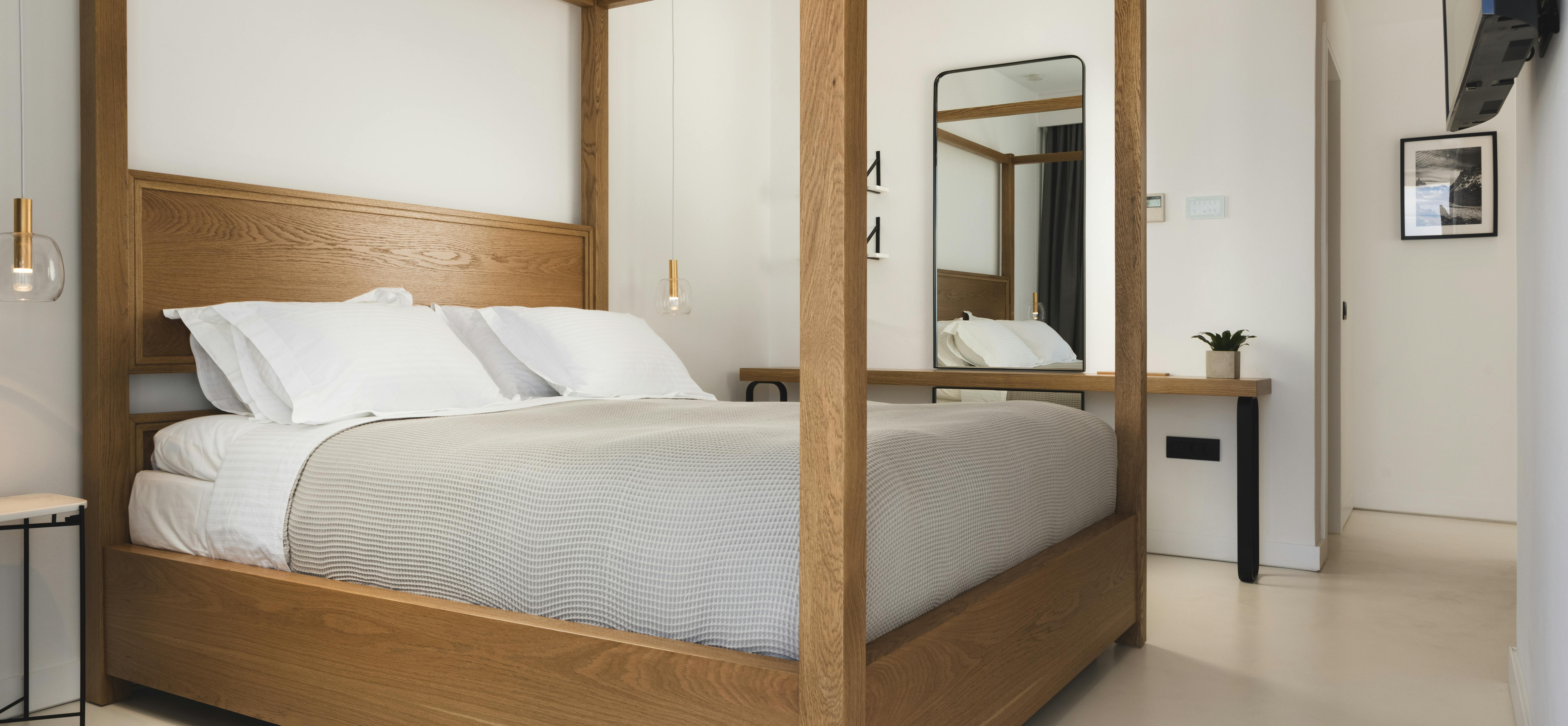 A contemporary bedroom features an elegant oak four-poster bed with crisp white linens, a large rounded mirror, modern wall sconces, and bright natural light flooding the space.