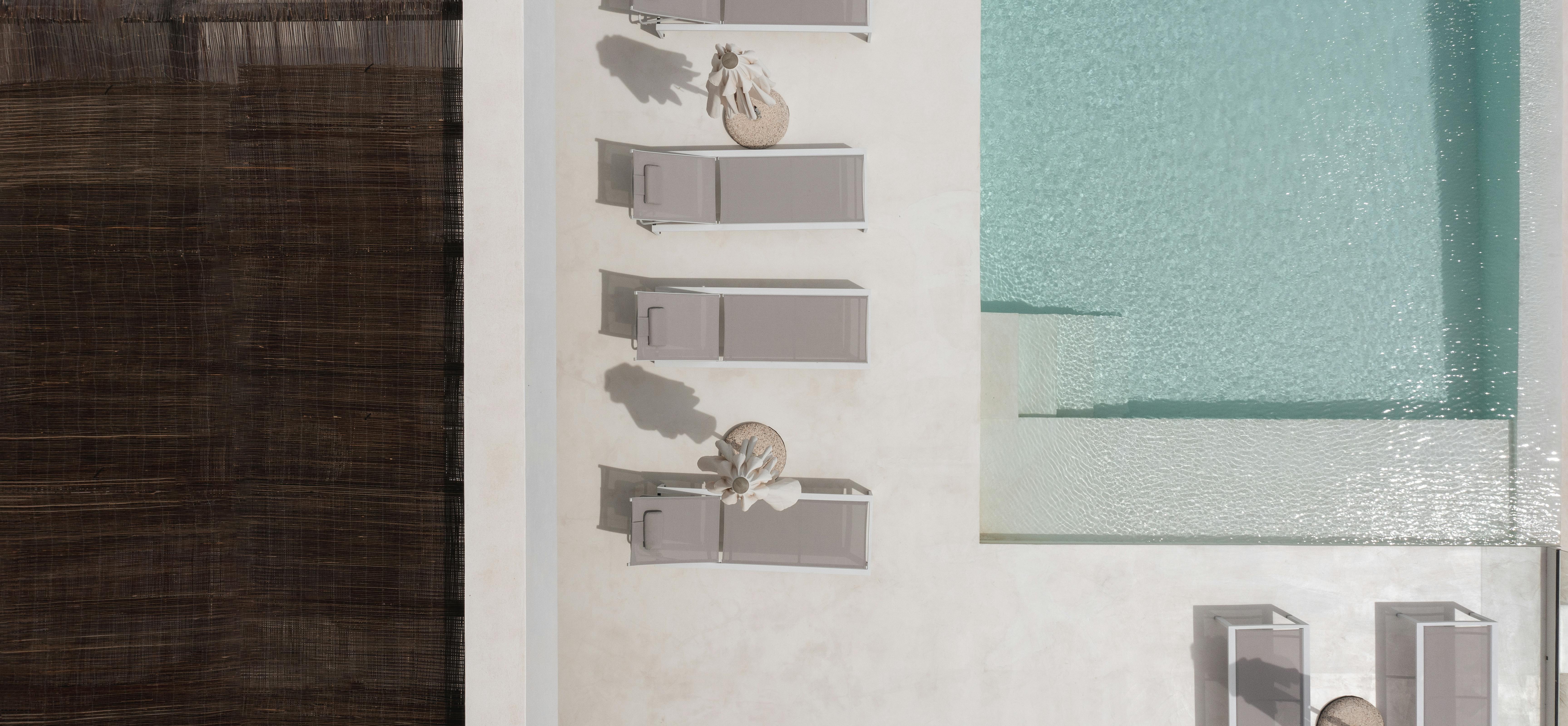 An overhead view shows modern grey loungers with white frames arranged in neat rows beside a turquoise infinity pool on white decking.