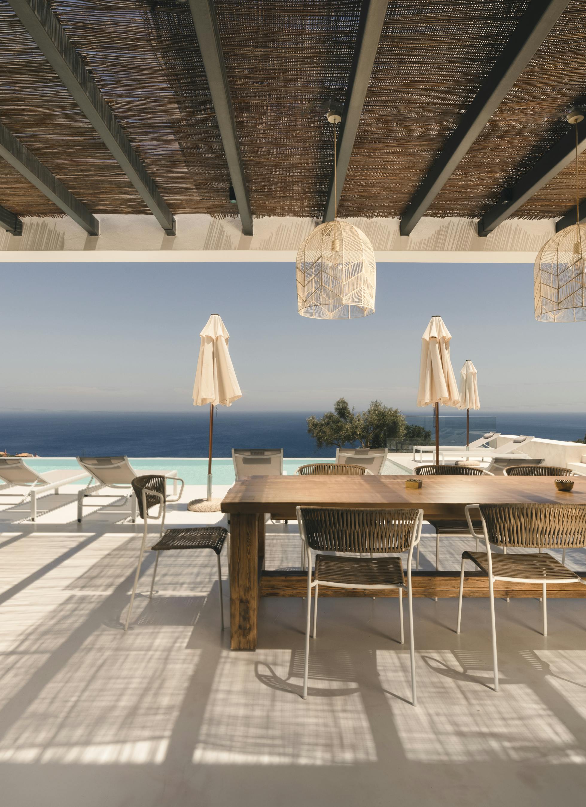 A covered outdoor terrace with traditional reed ceiling features a rustic wooden dining table, woven pendant lights, and dramatic views of the infinity pool and sea horizon.