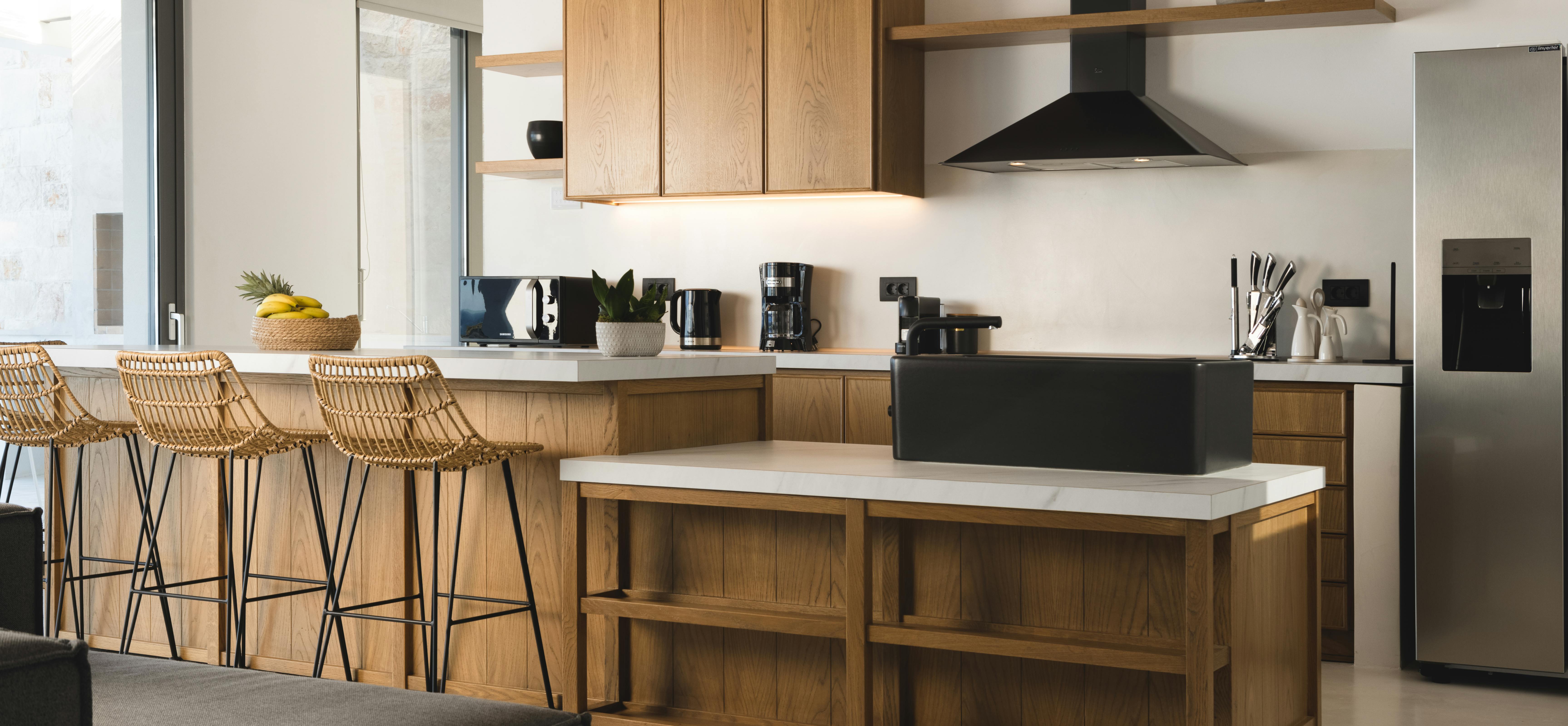 A contemporary kitchen showcases a substantial oak island with woven rattan bar stools, white countertops, black range hood, and pendant lights overhead.