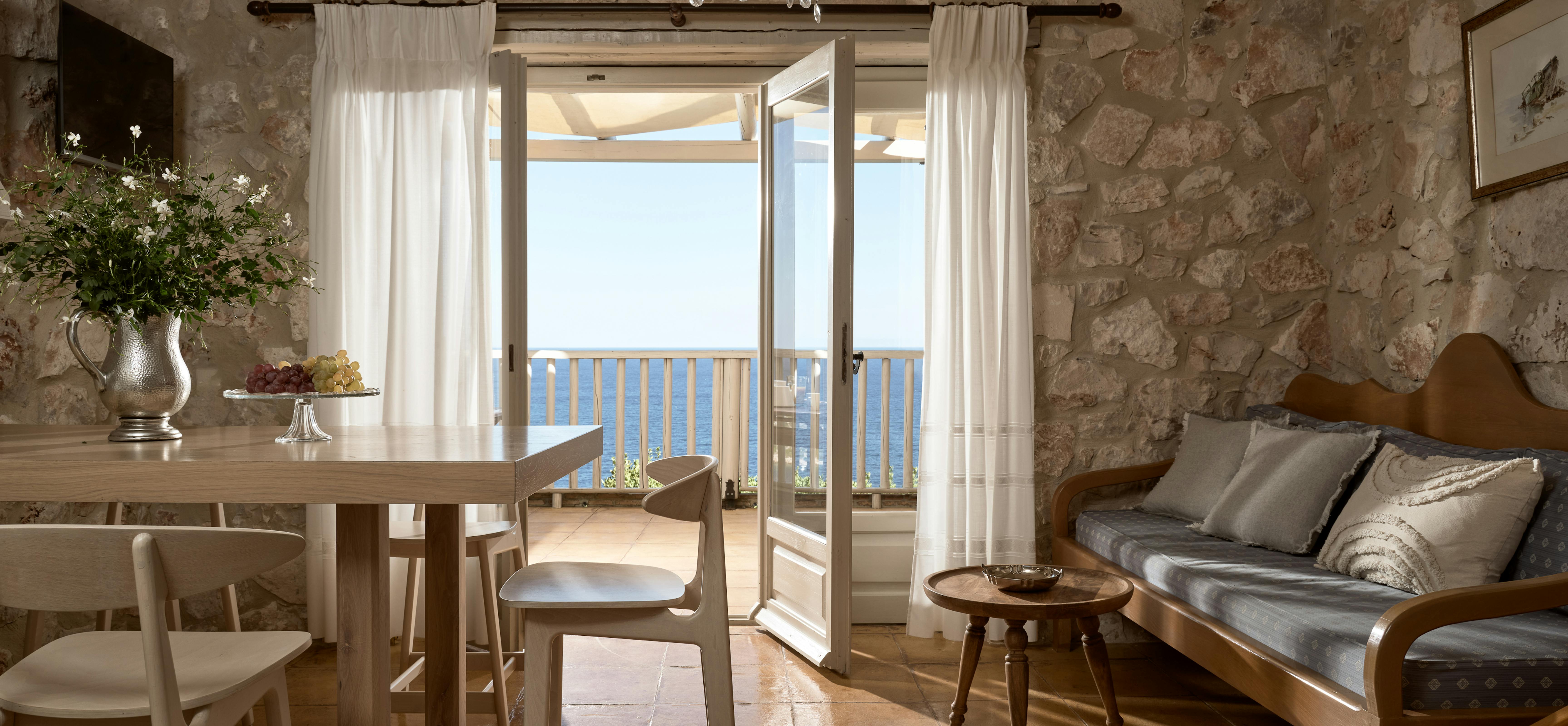 A rustic dining room with exposed stone walls and wooden beam ceiling features a crystal chandelier, white painted dining table with simple chairs, and French doors opening to a balcony with stunning sea views framed by flowing white curtains.