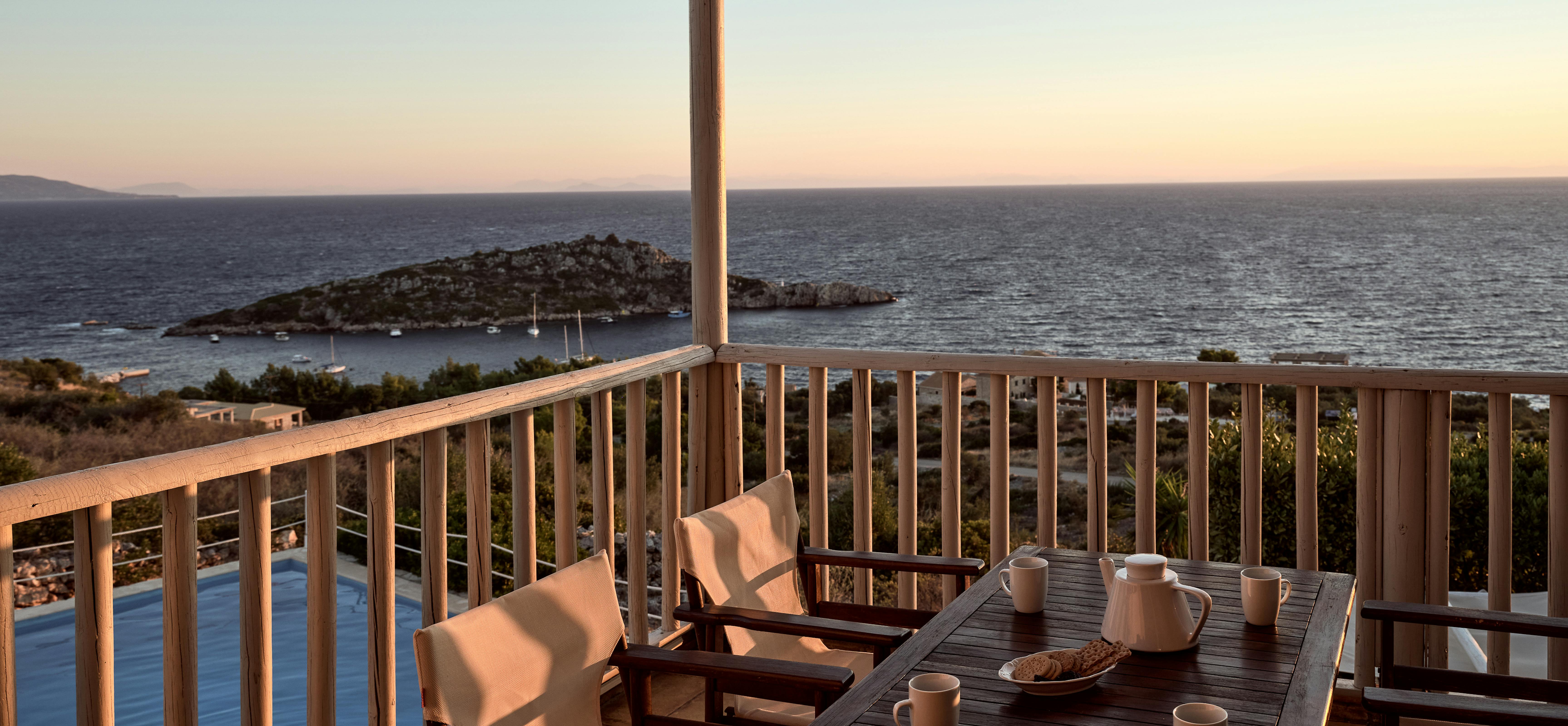 A covered terrace at golden hour features a dark wooden dining table set for breakfast with coffee and fresh pastries, overlooking a spectacular coastal panorama with rocky islets, anchored sailboats, and distant mountain ranges across the deep blue sea.